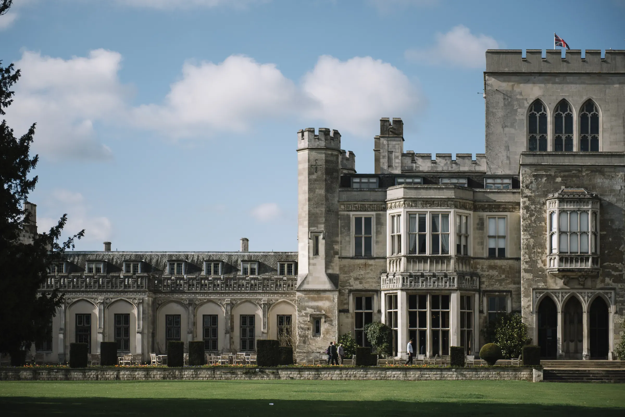 Historic stone building with ornate windows and turrets under a blue sky with clouds. People walking in the garden.