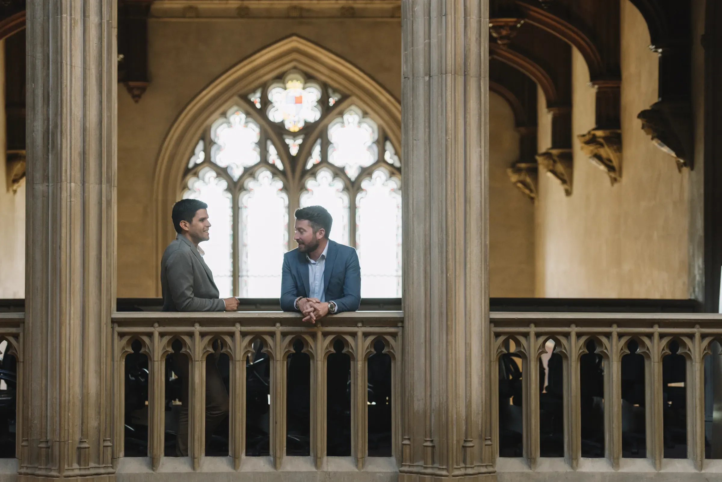 Two men in a historic building converse beside a stone railing, with arched windows and ornate architecture in the background.