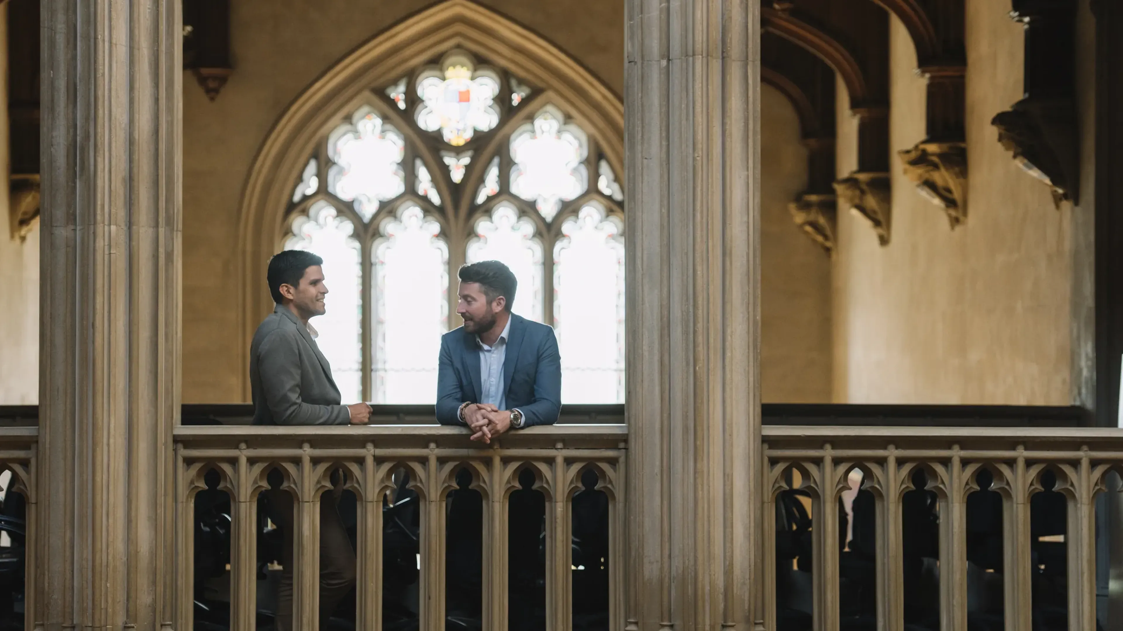 Two men in a historic building converse beside a stone railing, with arched windows and ornate architecture in the background.