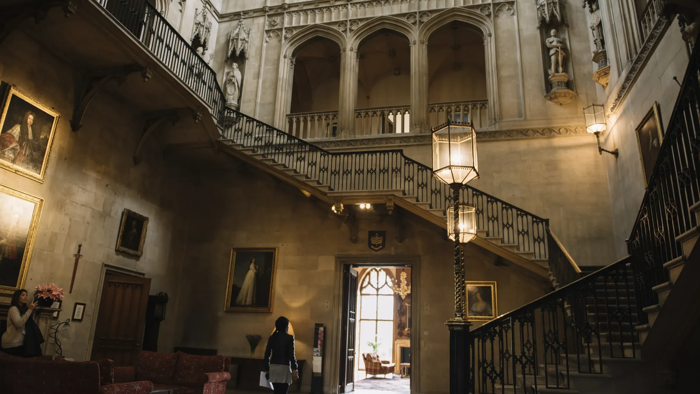 A grand, historic hall with portraits, ornate staircases, and soft lighting. A person walks through, admiring the architecture.