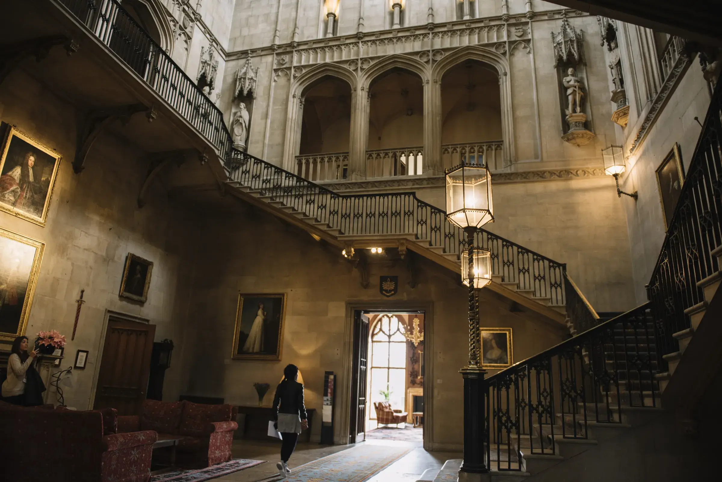 A grand, historic hall with portraits, ornate staircases, and soft lighting. A person walks through, admiring the architecture.
