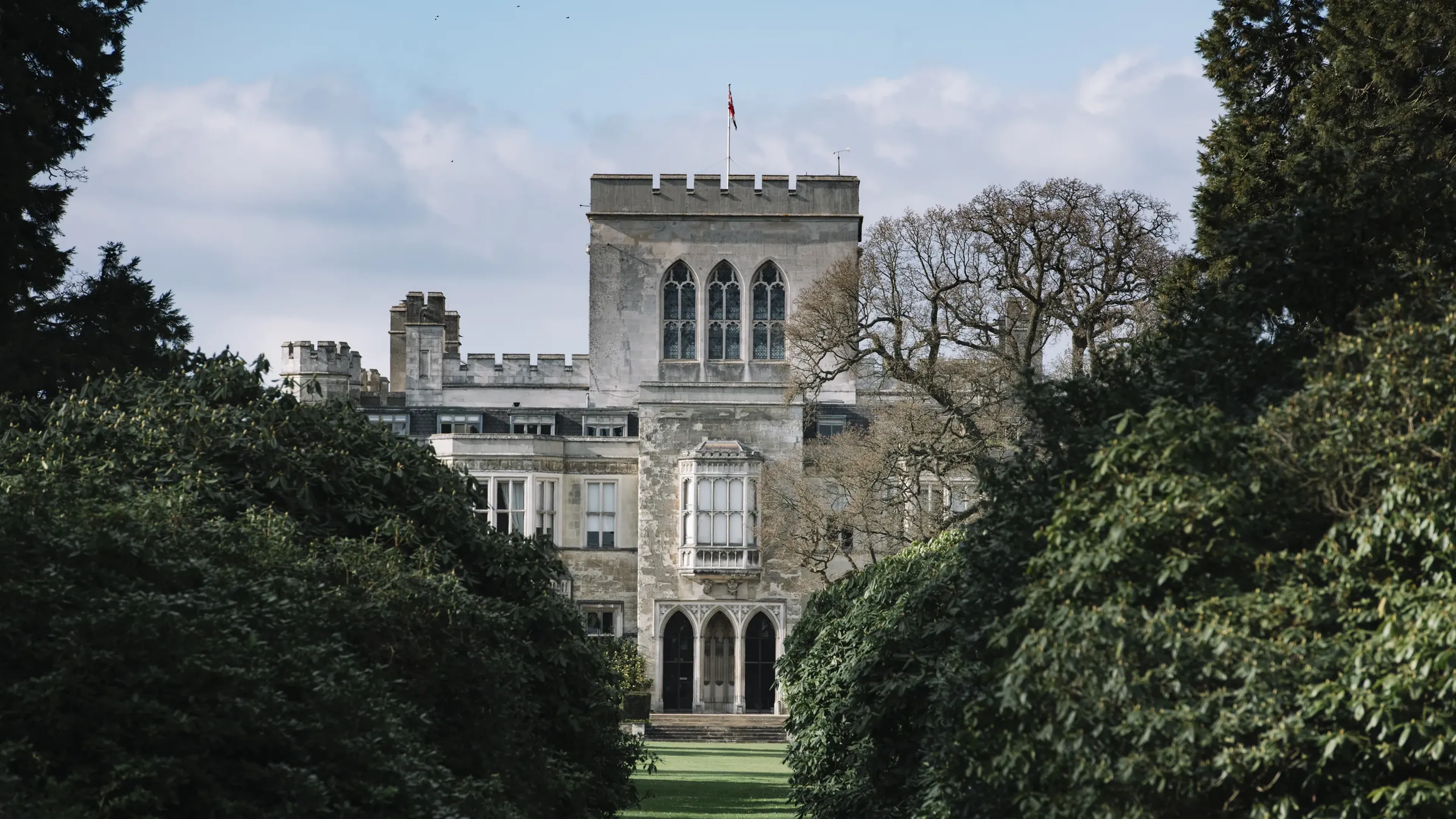 A historic stone building with arched windows and a central tower, surrounded by lush greenery and trees under a blue sky.