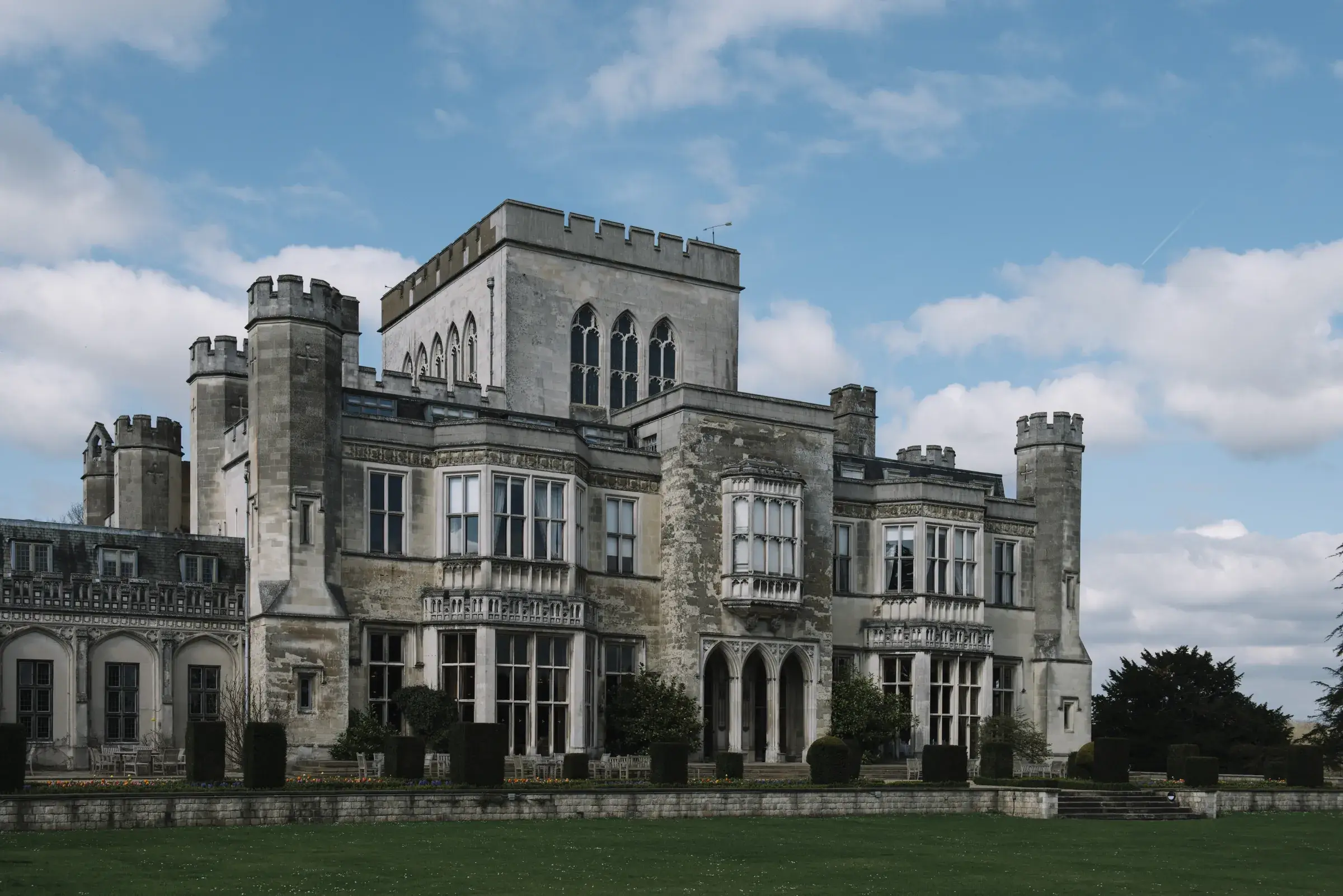 Large, historic stone building with turrets and arched windows, set against a blue sky with scattered clouds.