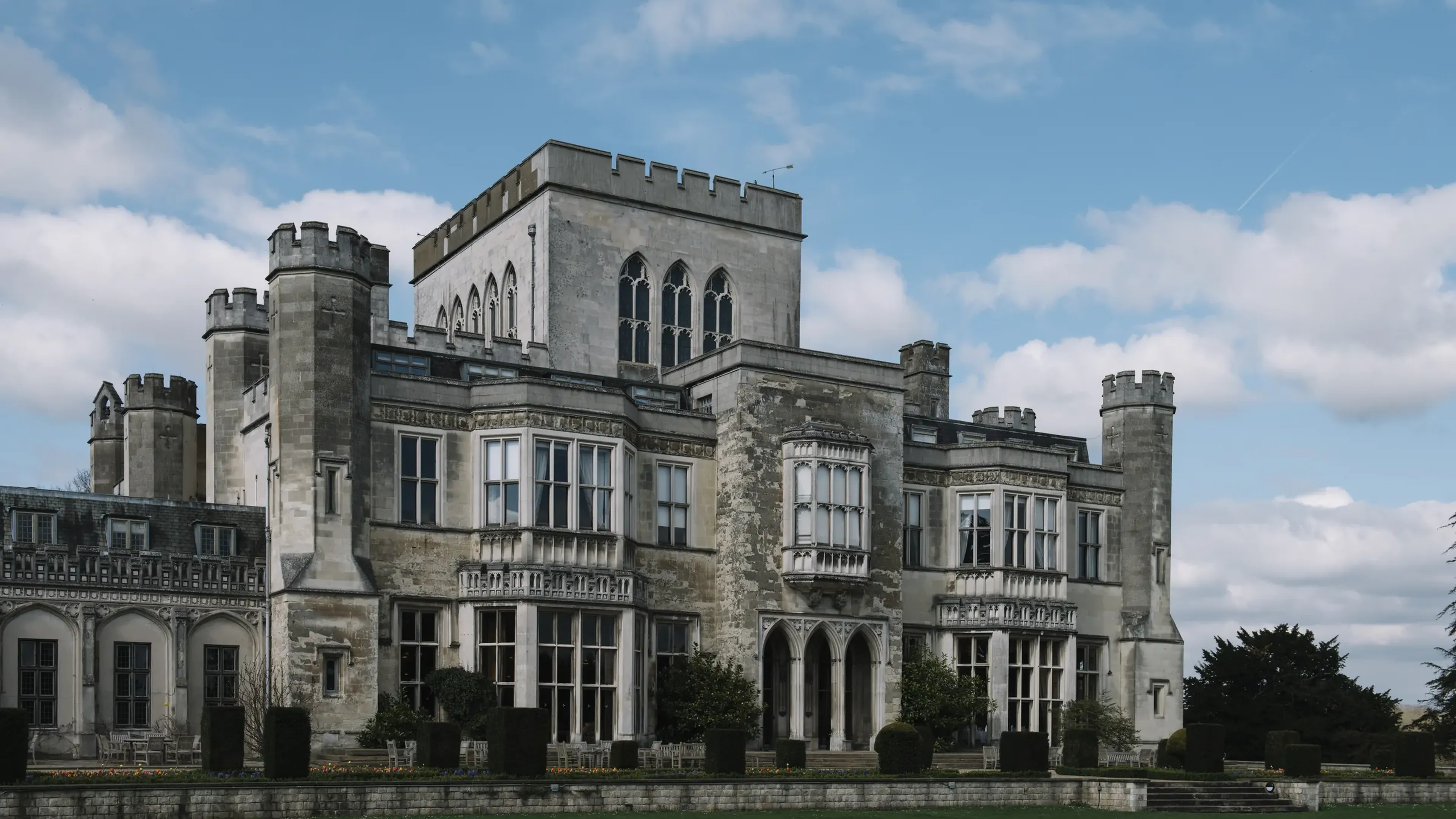 Large, historic stone building with turrets and arched windows, set against a blue sky with scattered clouds.