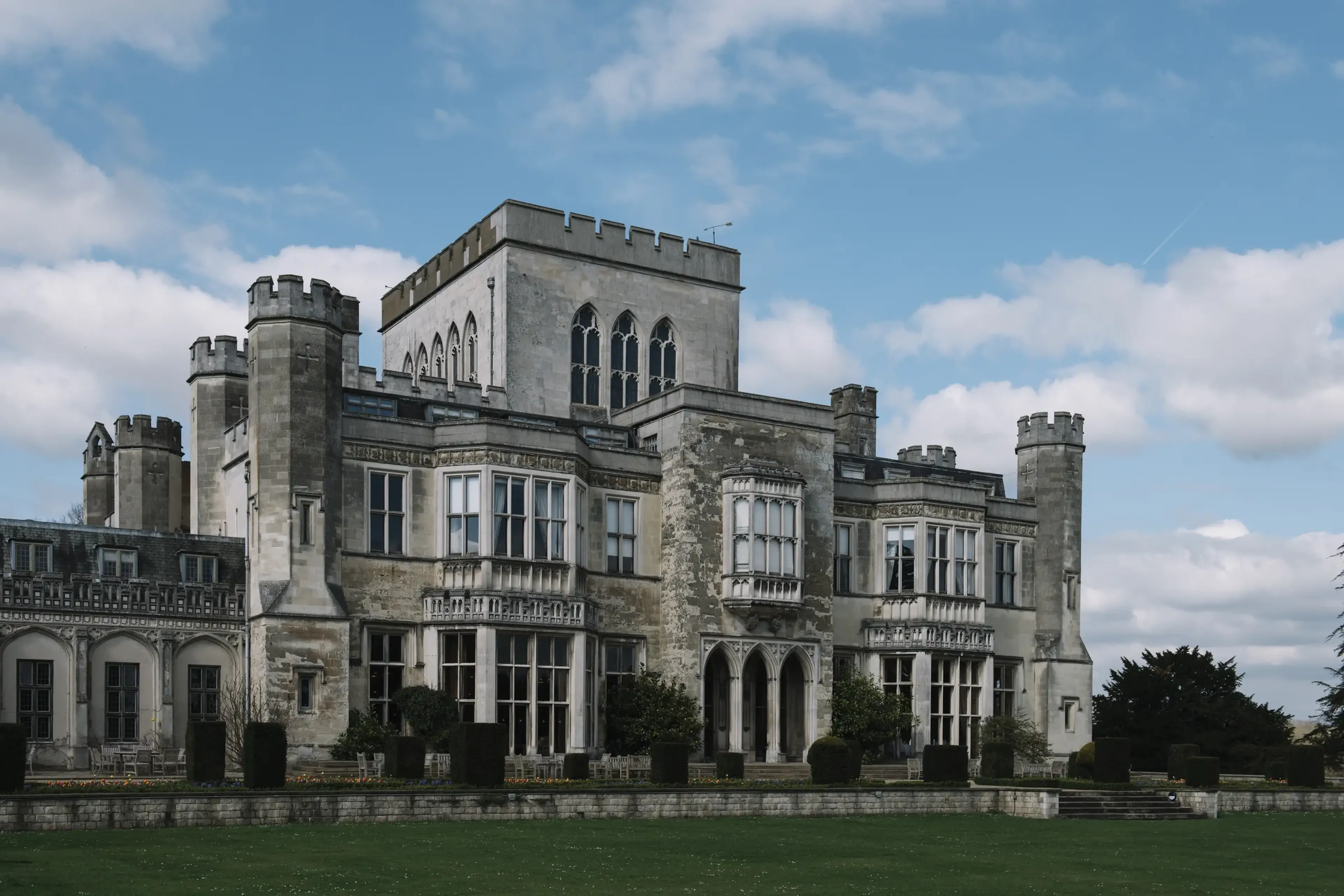 Large, historic stone building with turrets and arched windows, set against a blue sky with scattered clouds.