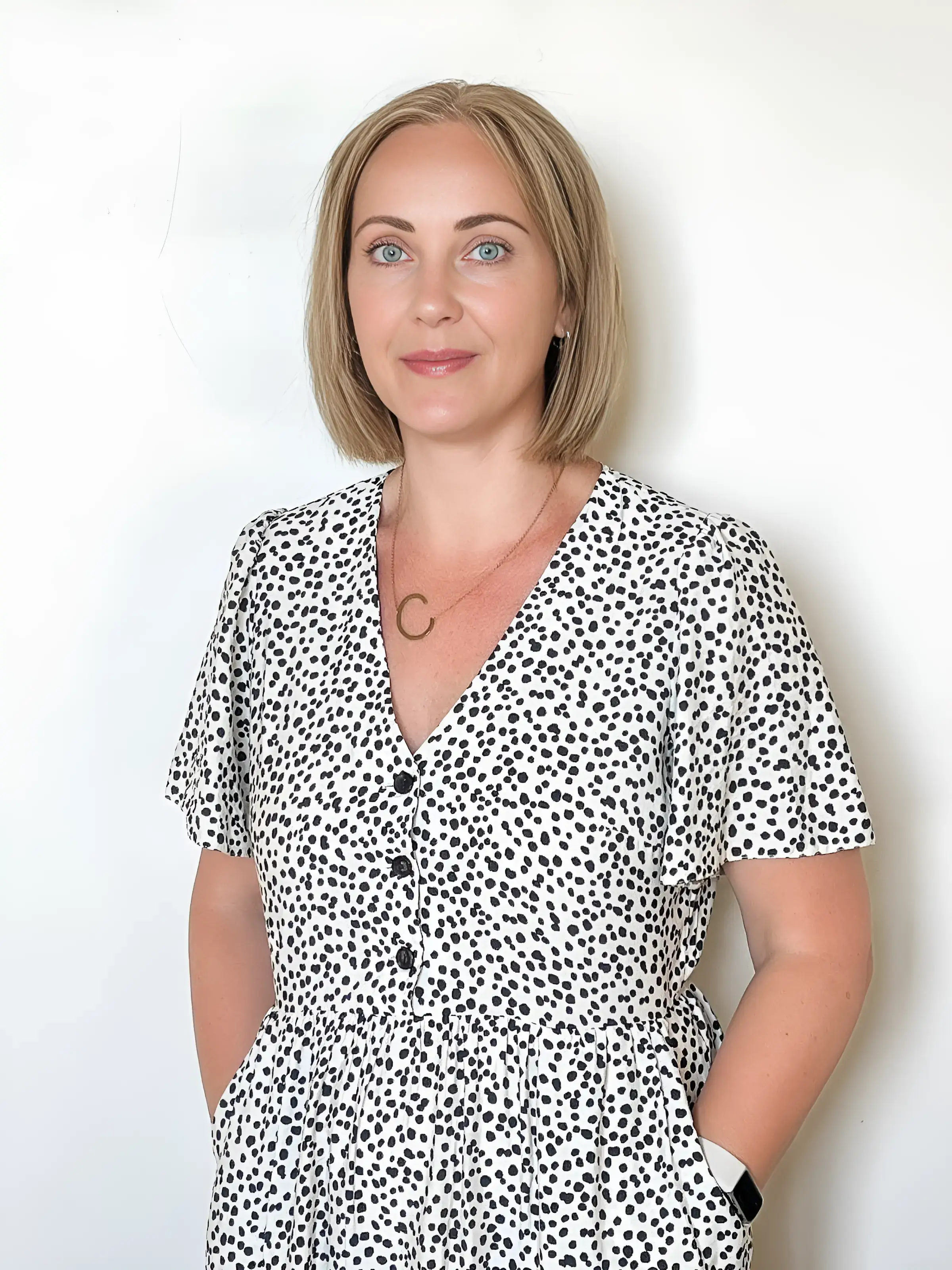 A woman in a polka dot dress stands against a white background, smiling.