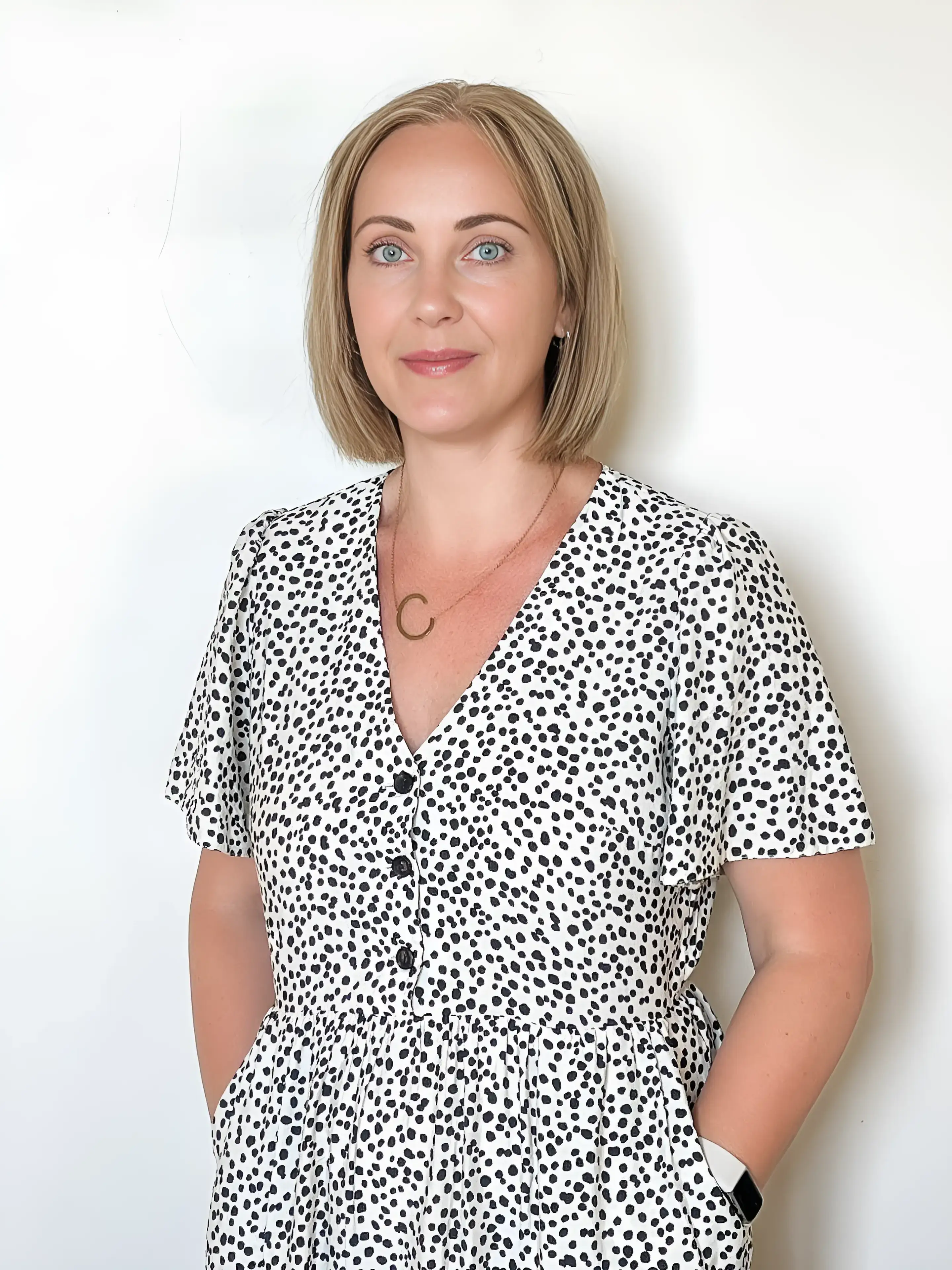 A woman in a polka dot dress stands against a white background, smiling.