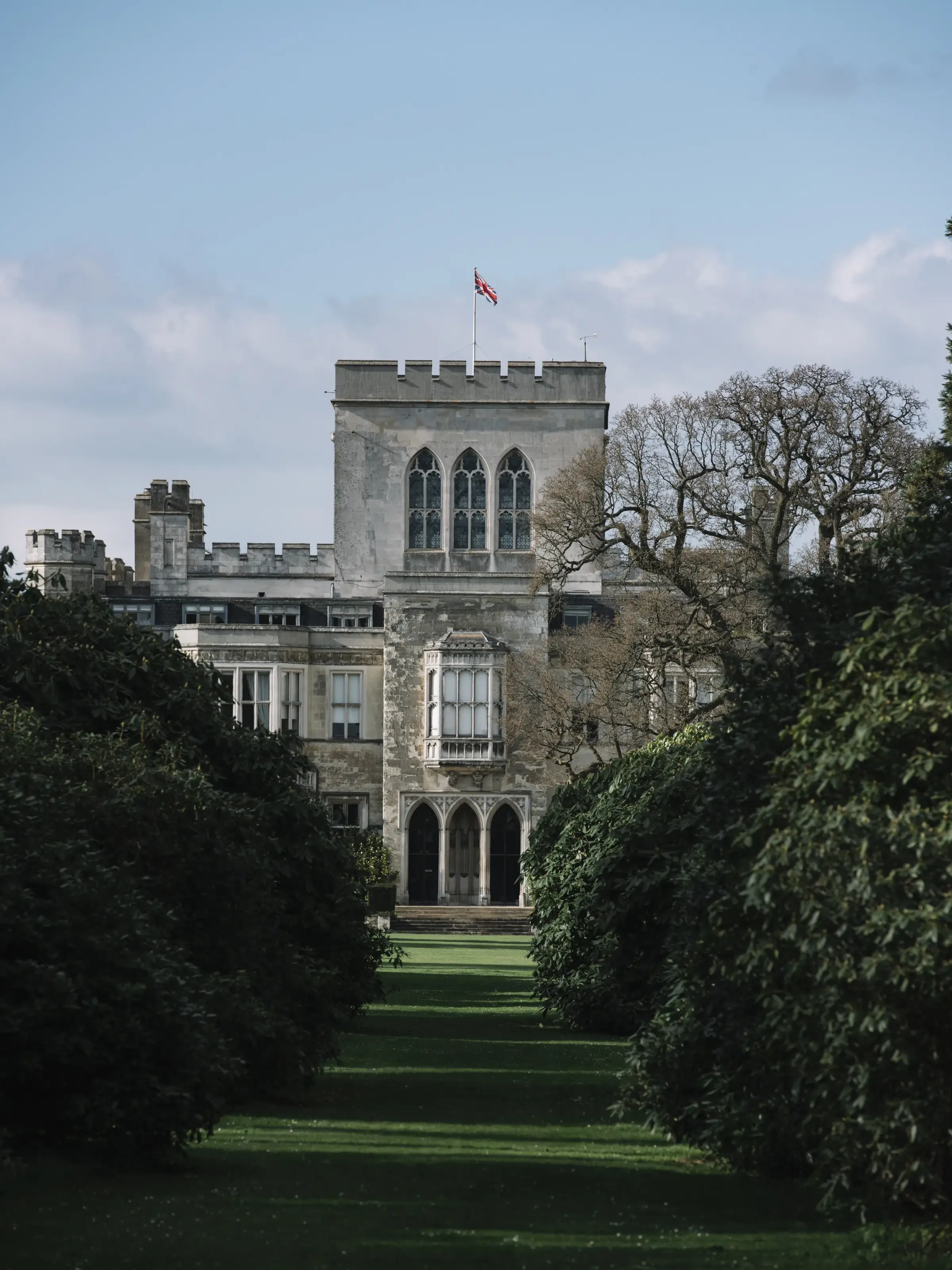 Stone castle with a Union Jack flag, large arched windows, and surrounded by lush greenery, under a clear blue sky.
