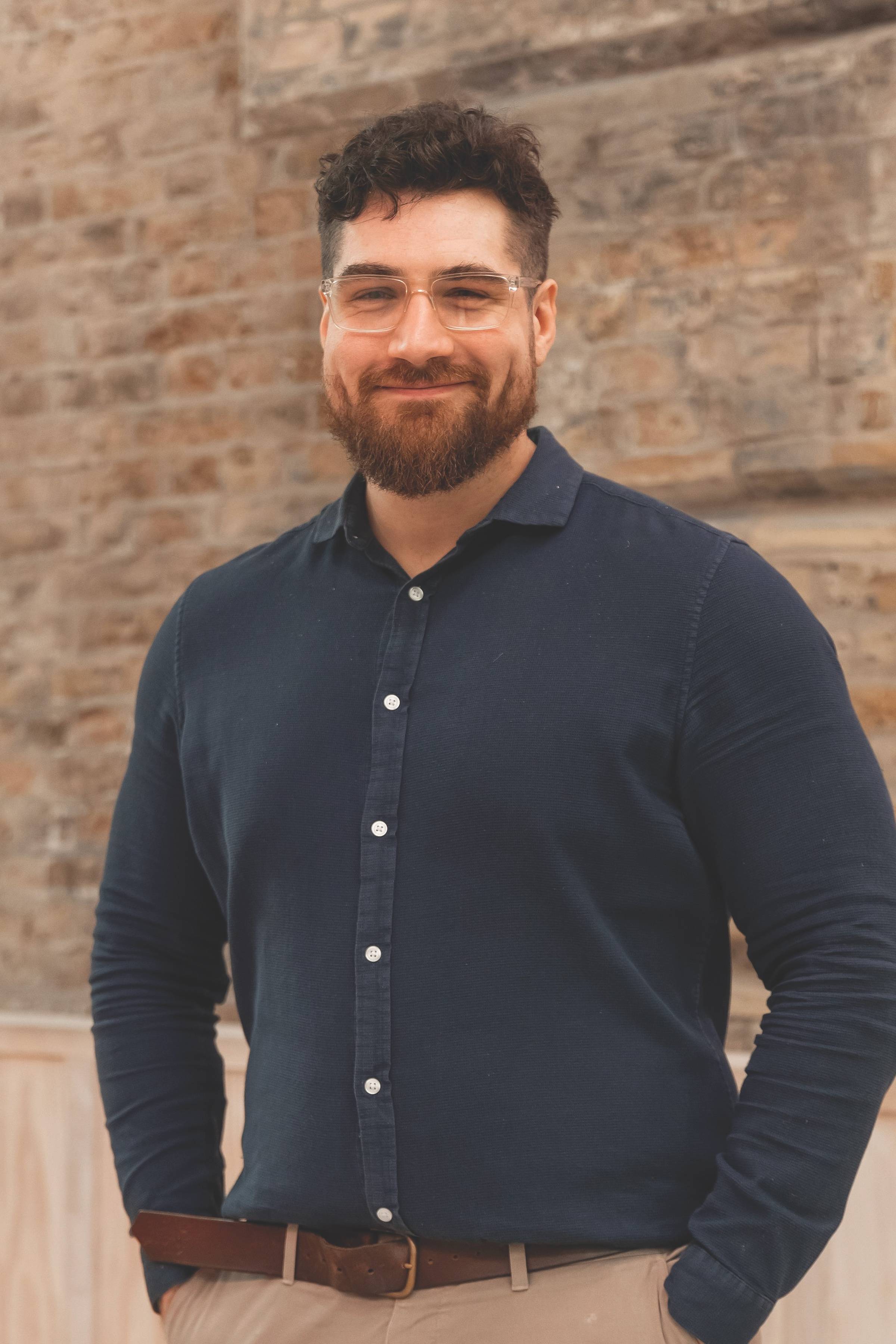 Smiling man with glasses in a navy shirt and khaki pants stands against a brick wall background, hands in pockets.
