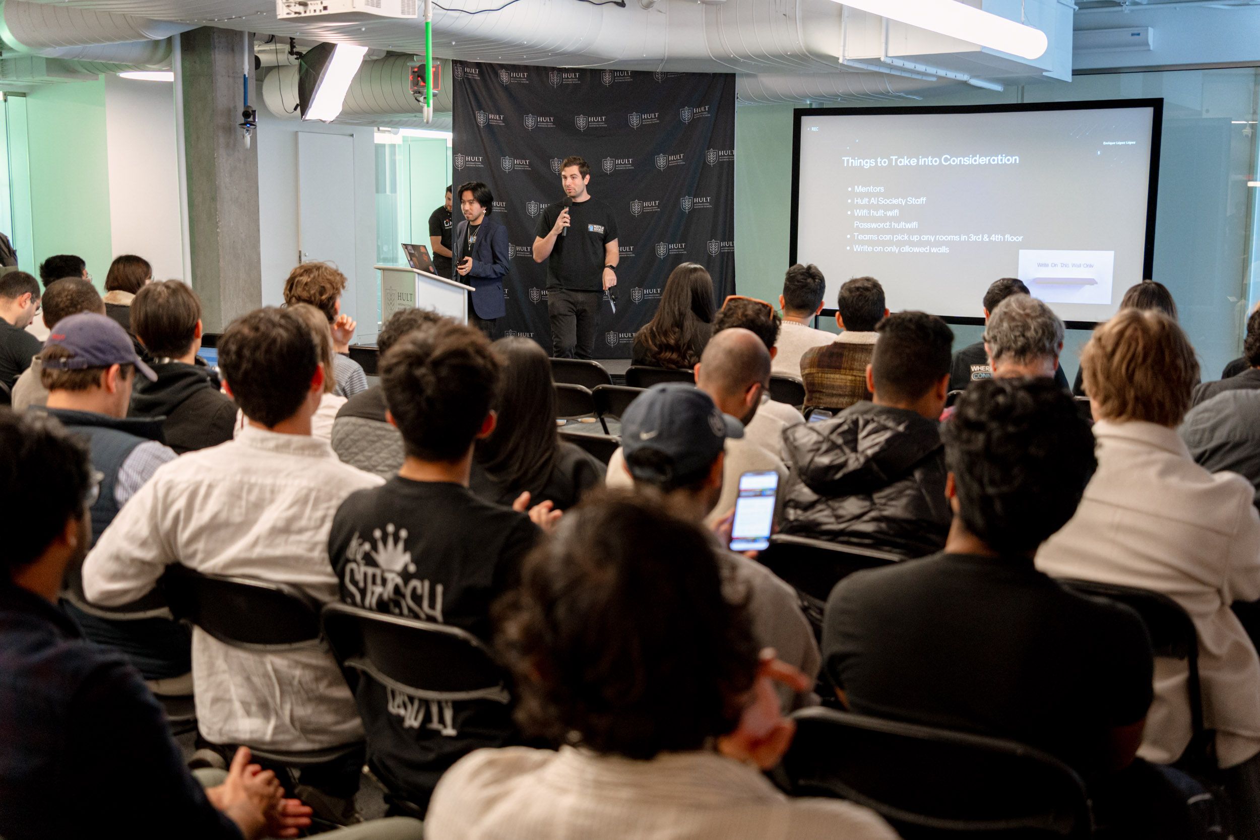 Audience seated in a conference room listens to two presenters at the front, with a presentation slide displayed on a screen.