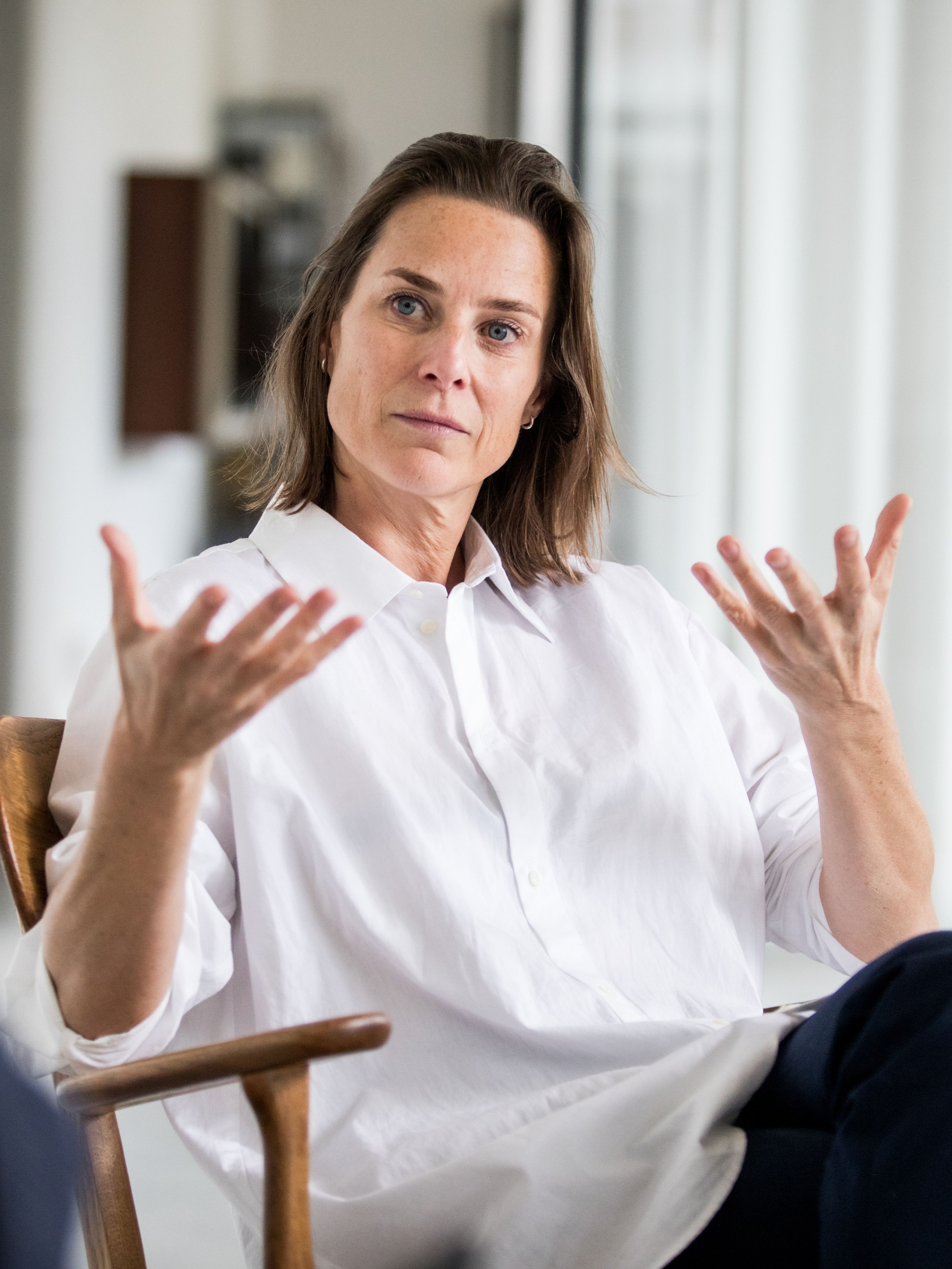 A woman in a white shirt gestures expressively with her hands while sitting on a wooden chair, engaged in conversation.