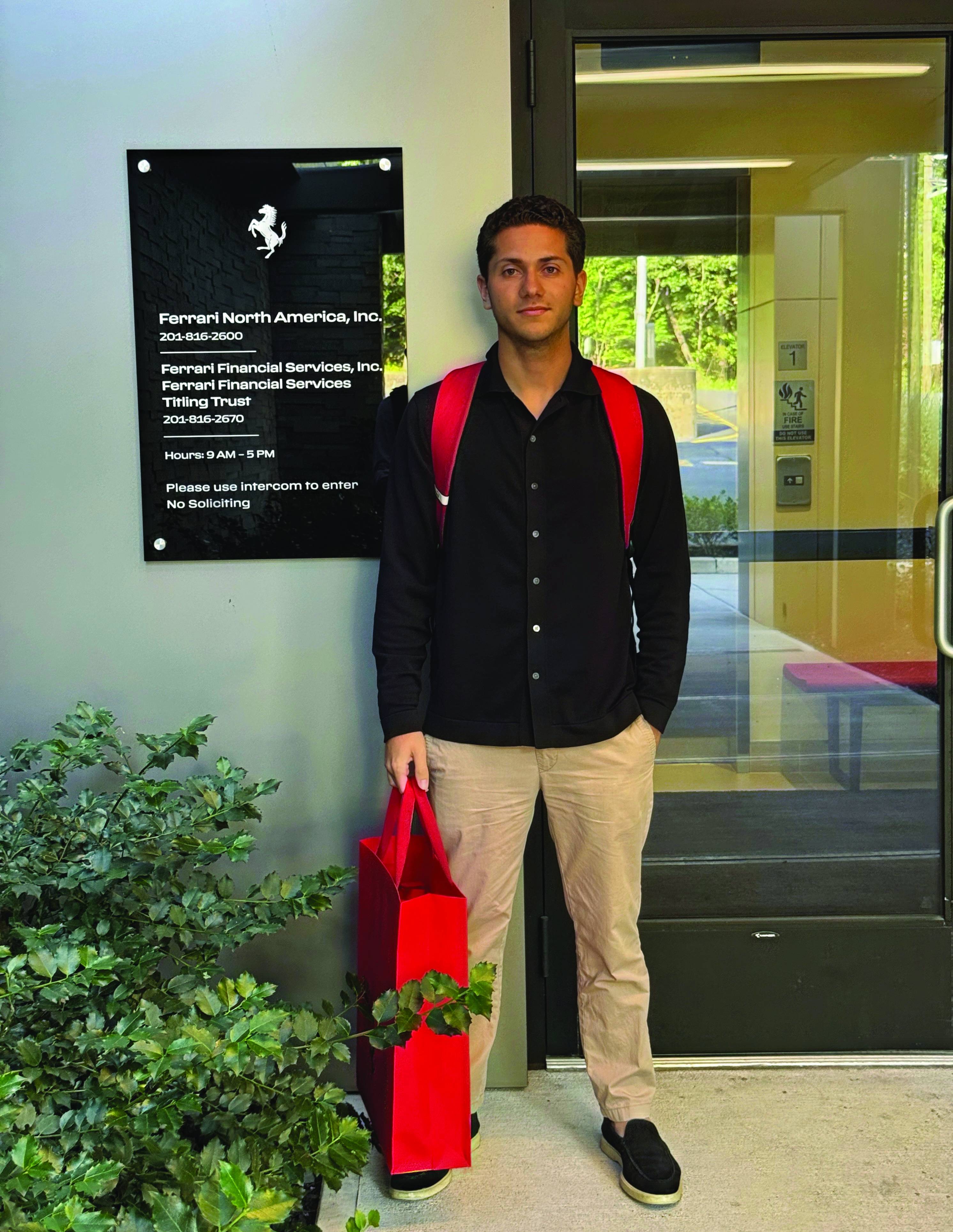 Man with a red backpack stands outside Ferrari North America office, holding a red shopping bag. Office signage visible beside him.