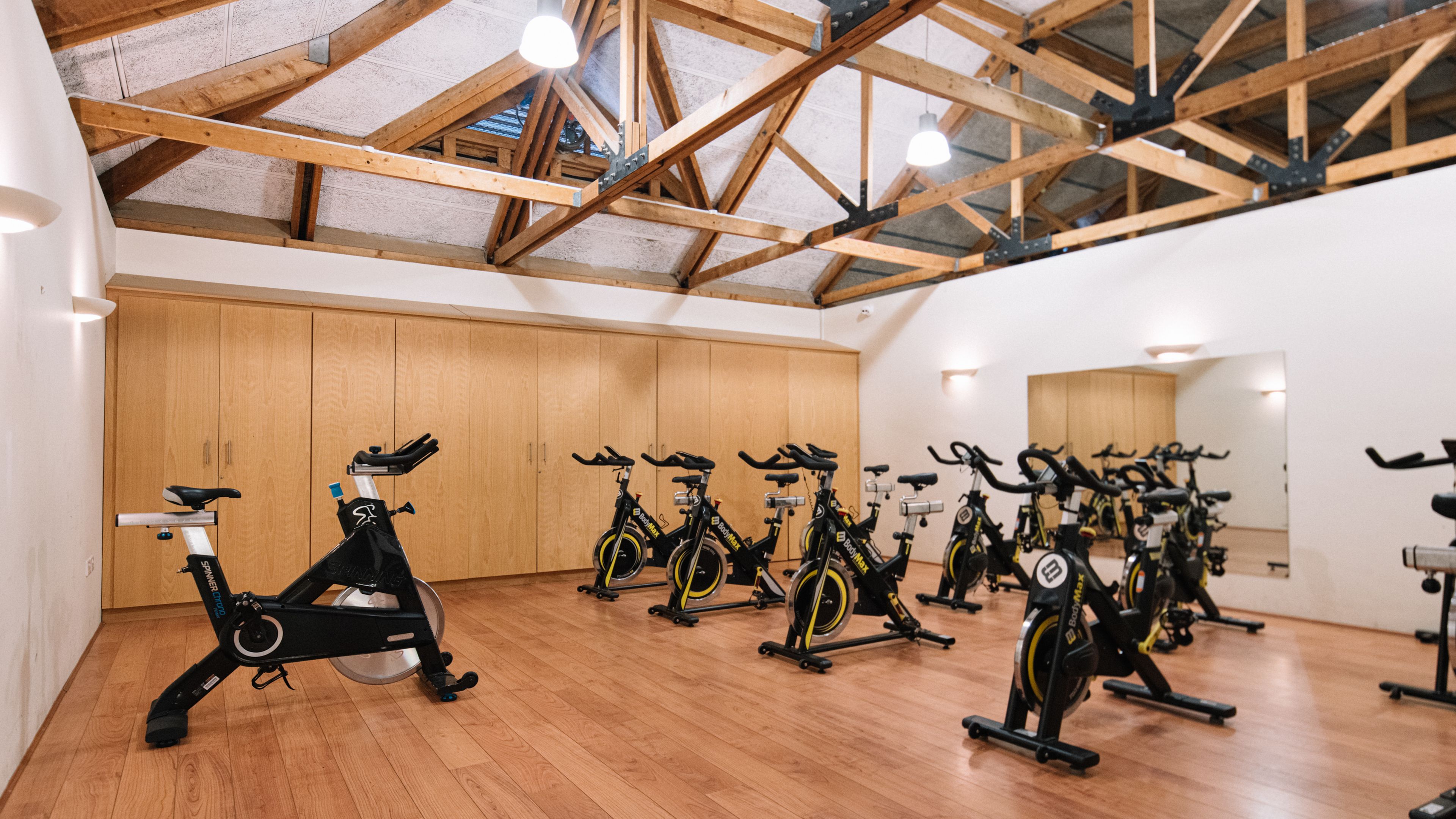 A gym room with several stationary bikes on a wooden floor under exposed wooden beams.