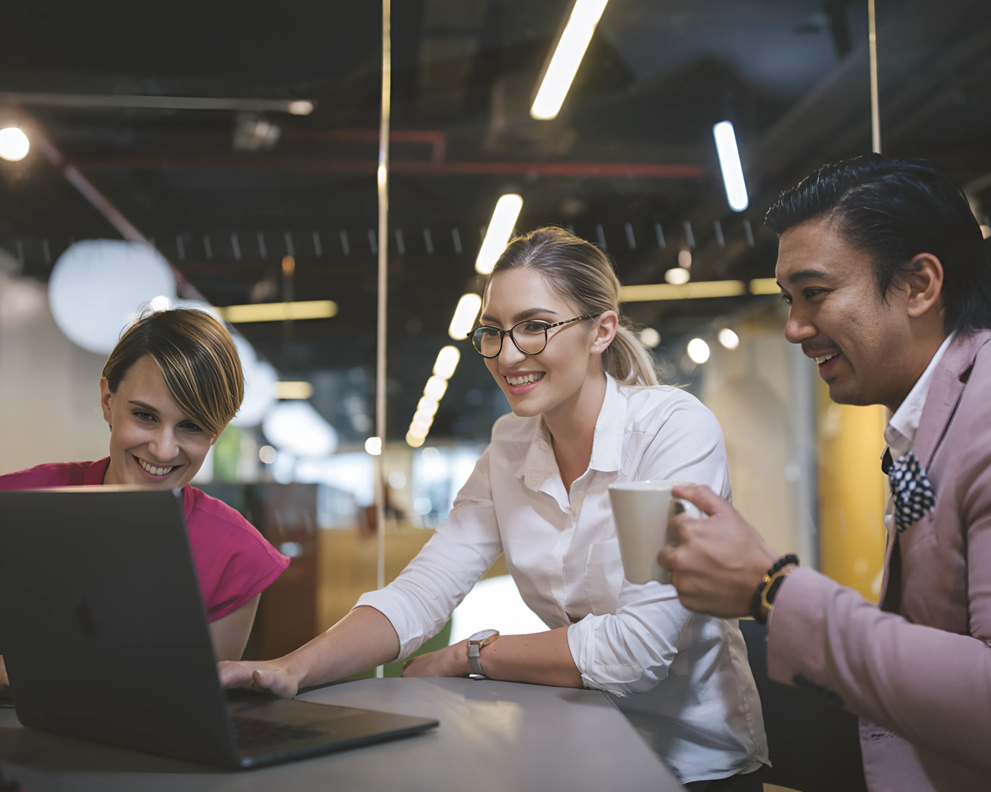 Three people smiling at a laptop screen in an office setting.