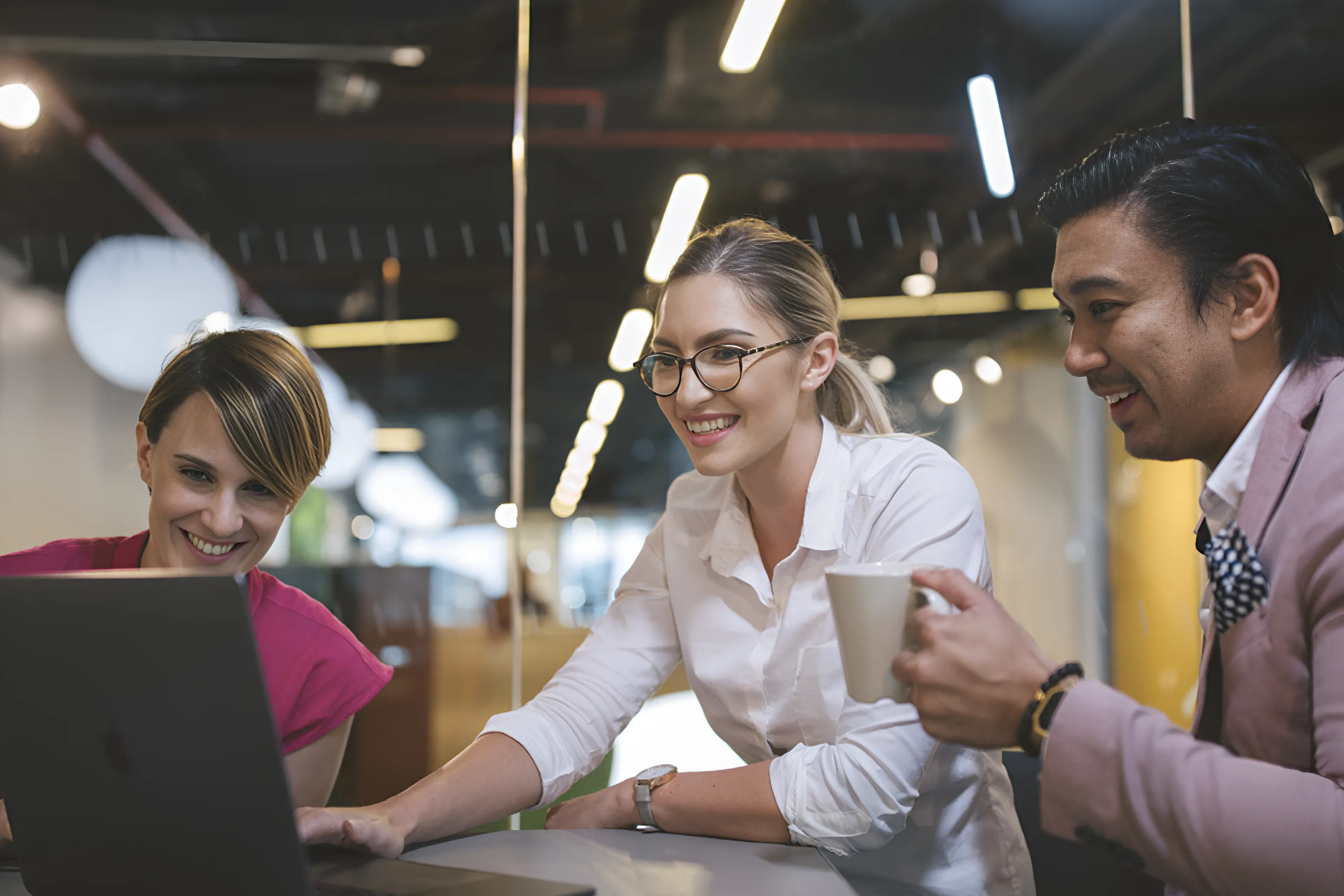 Three people smiling at a laptop screen in an office setting.