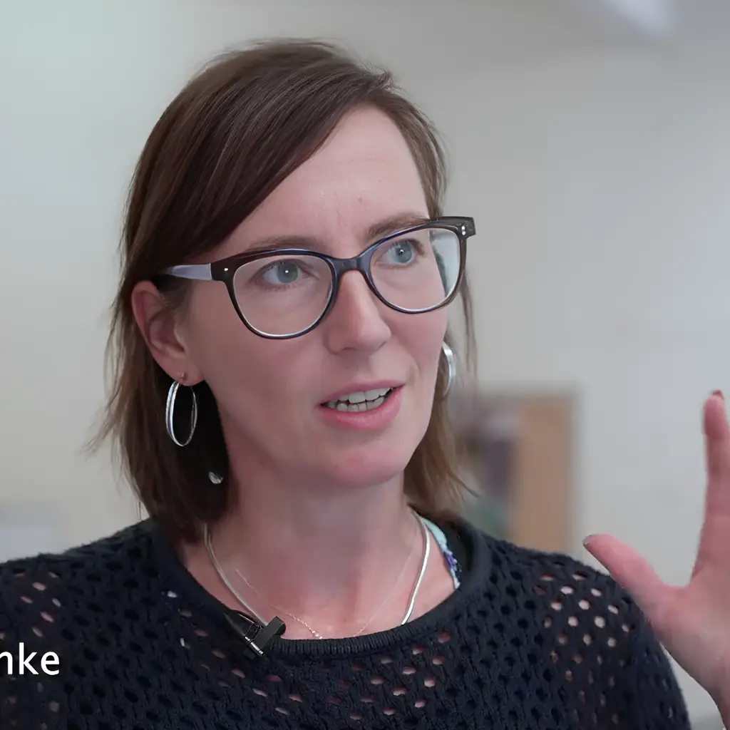 A woman with glasses speaks, gesturing with her hands, wearing a black top.