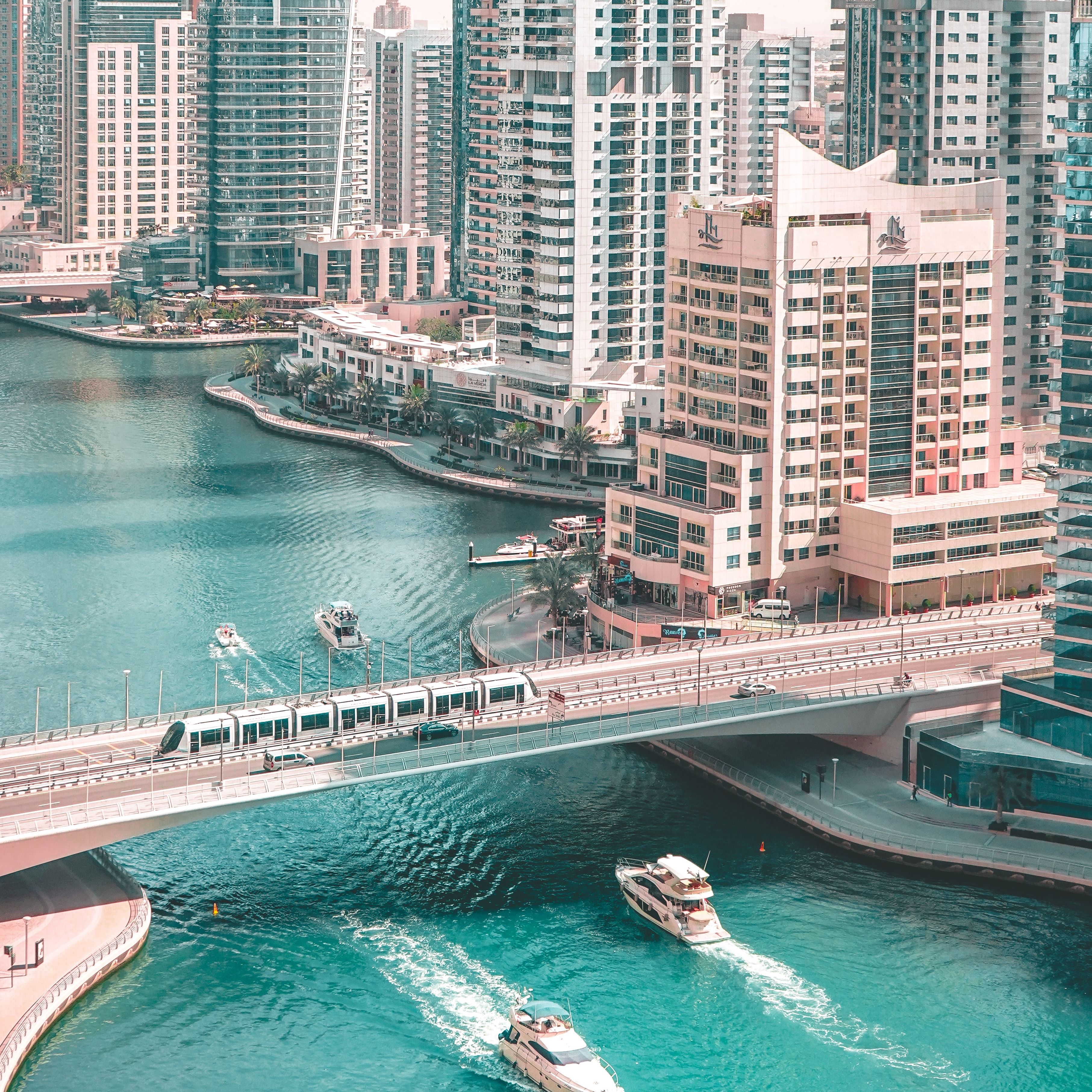 Aerial view of a cityscape with tall buildings, a river, two boats, and a bridge crossing the water.
