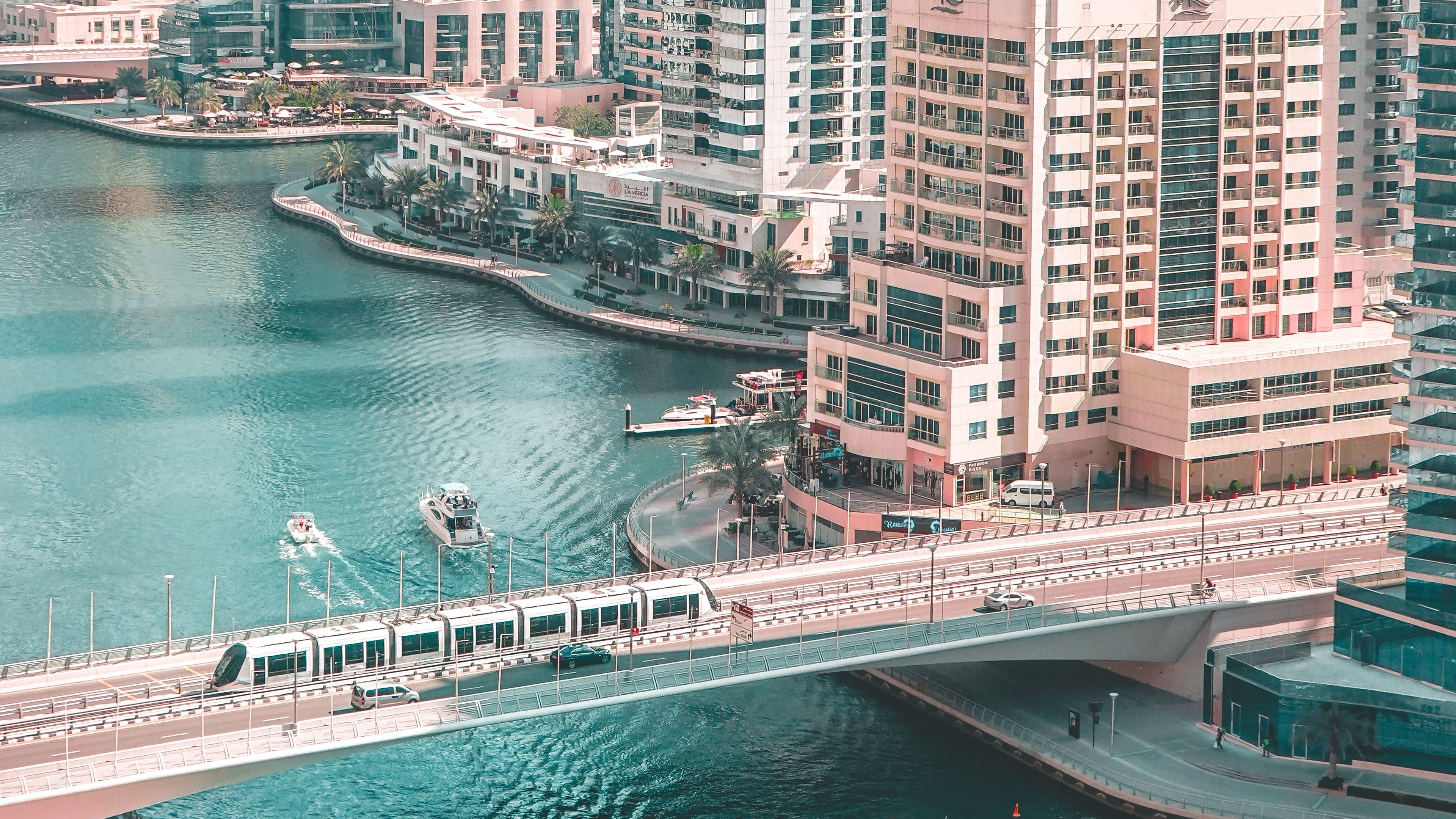 Aerial view of a cityscape with tall buildings, a river, two boats, and a bridge crossing the water.