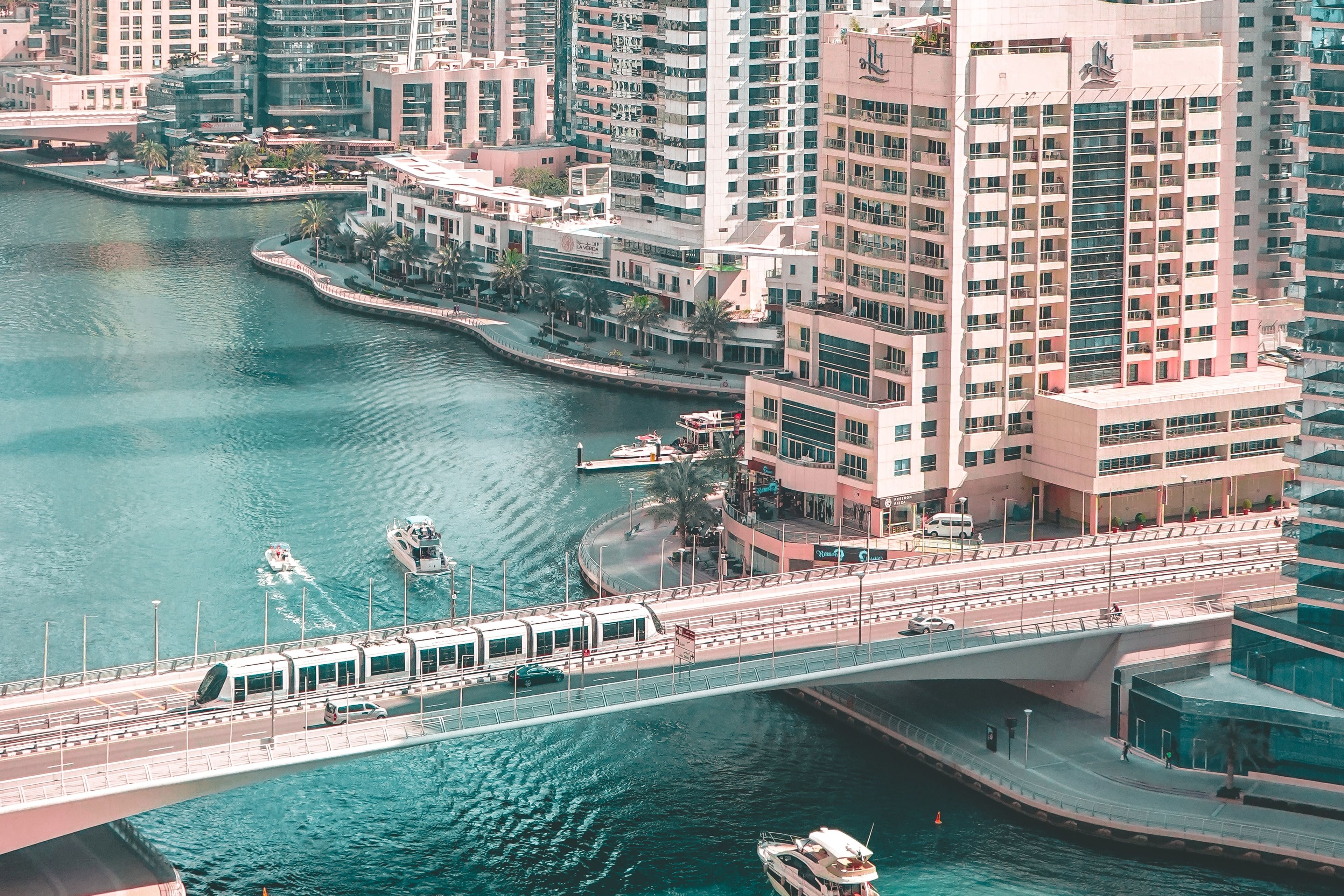 Aerial view of a cityscape with tall buildings, a river, two boats, and a bridge crossing the water.