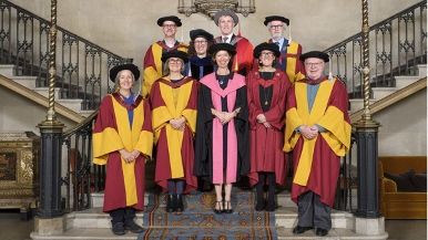 A group of nine people in academic regalia pose on a staircase, with ornate railings and a patterned carpet visible.