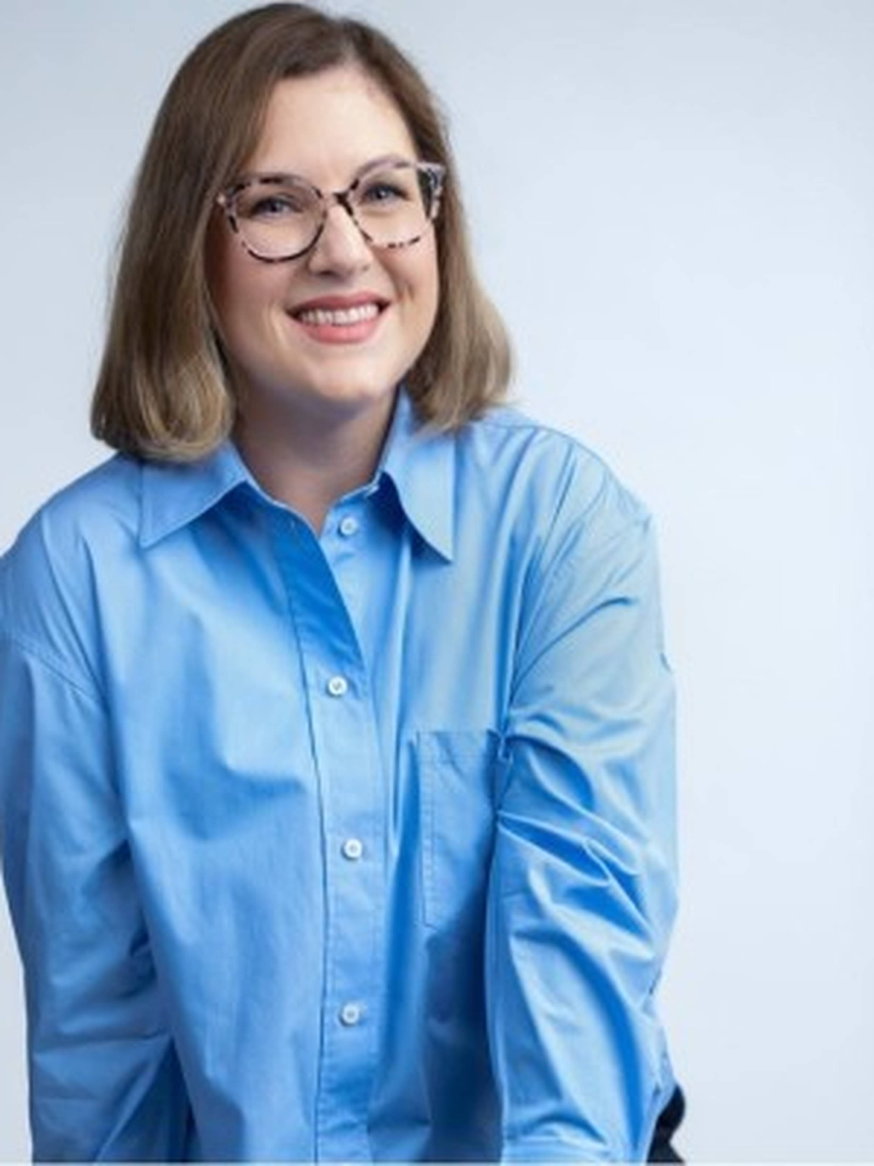 Smiling woman with glasses wearing a blue shirt, standing against a light background.