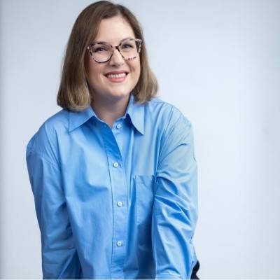 Smiling woman with glasses wearing a blue shirt, standing against a light background.