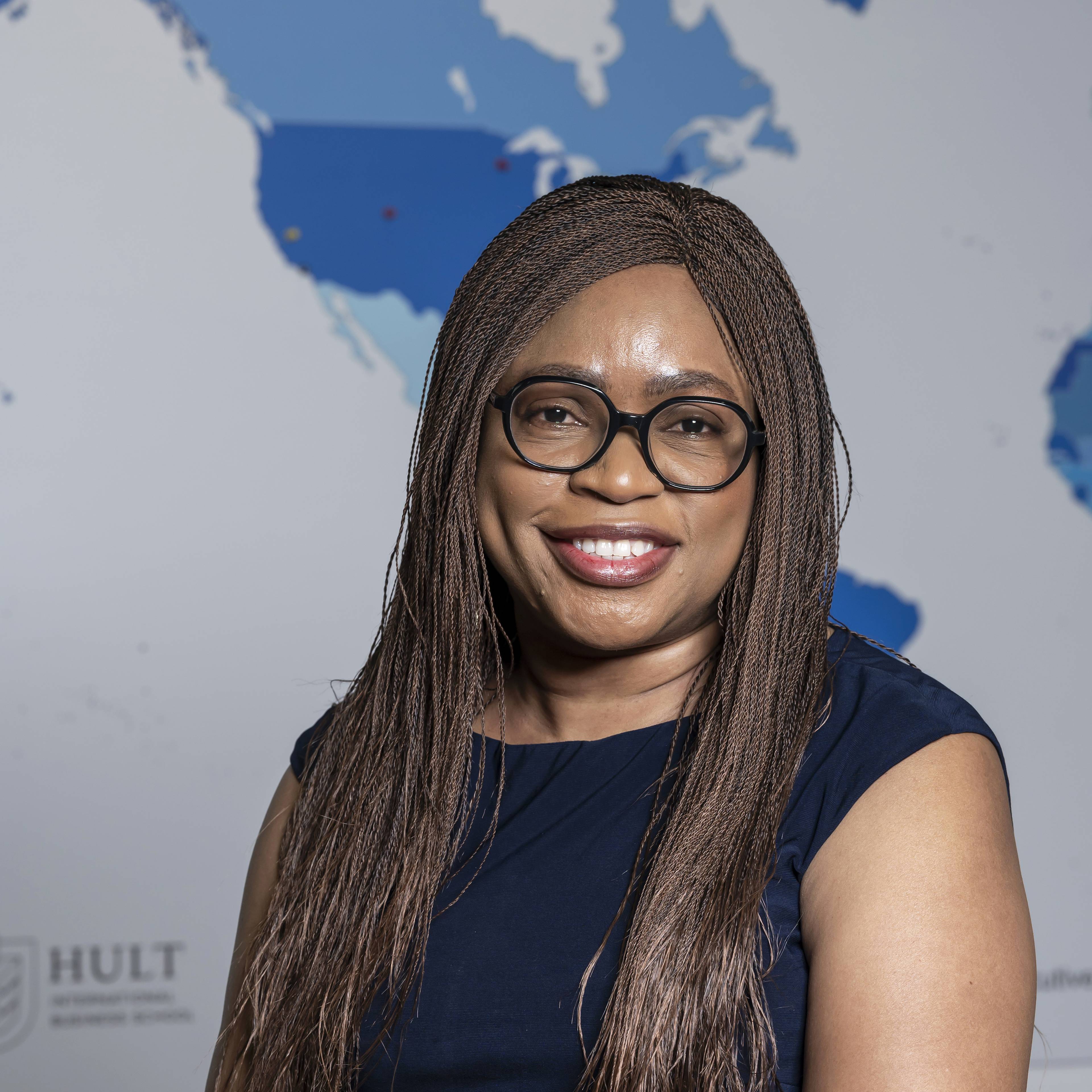 A woman in glasses and a navy dress smiles in front of a world map.