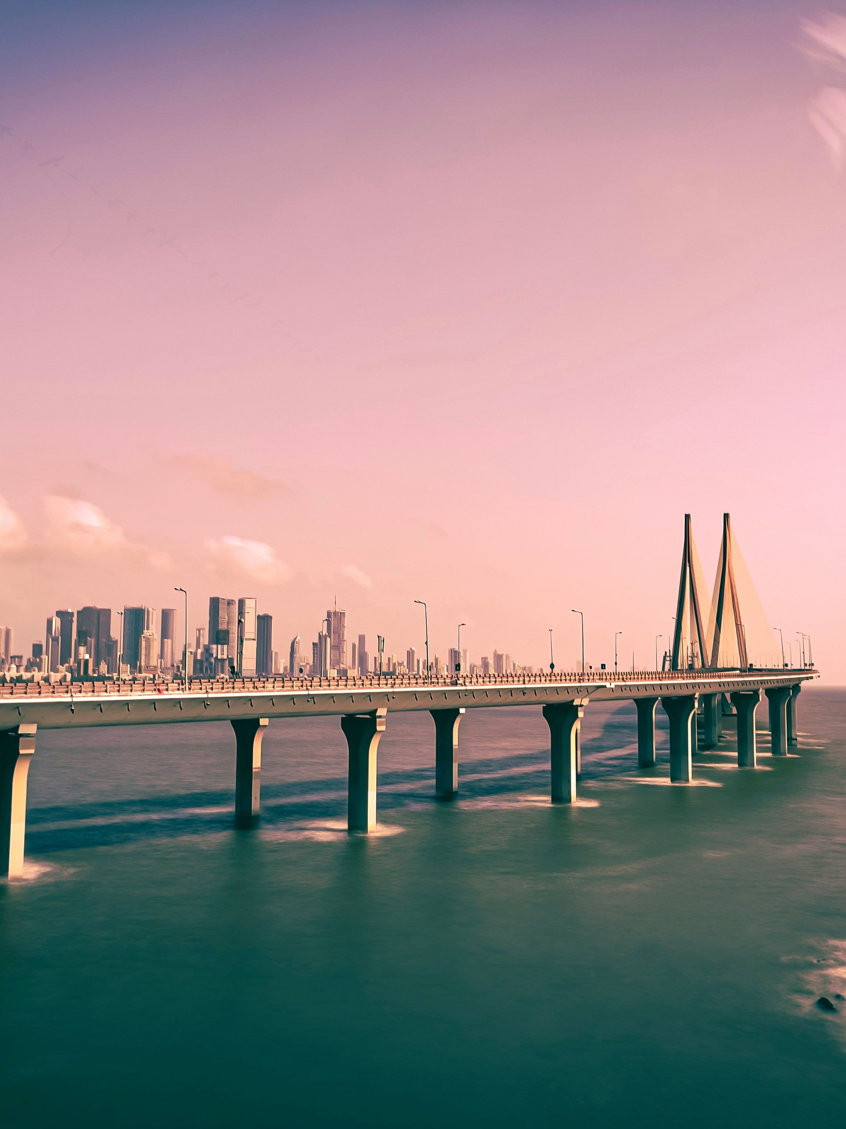 A bridge extends over the ocean towards a city skyline under a pink-tinted sky.