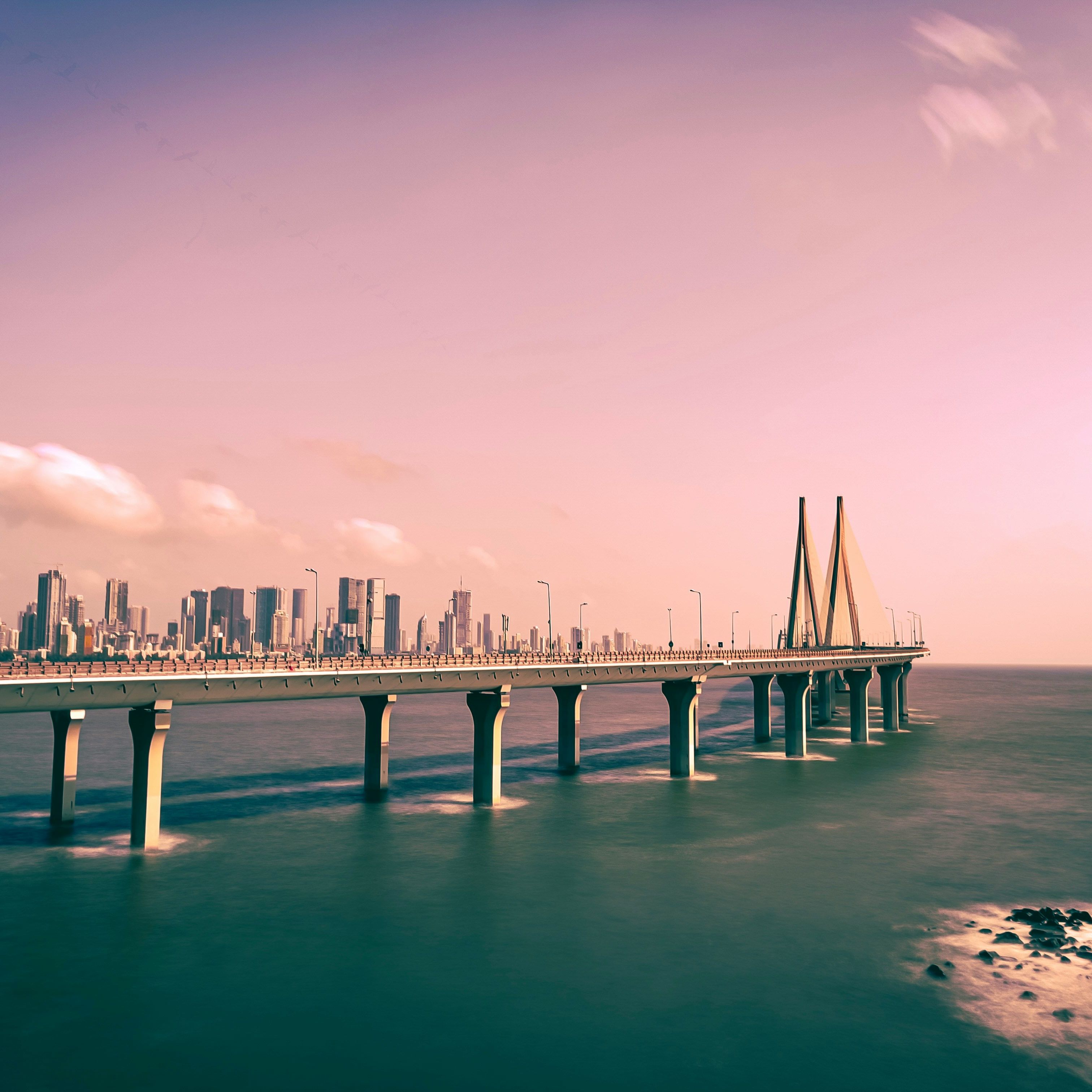 A bridge extends over the ocean towards a city skyline under a pink-tinted sky.