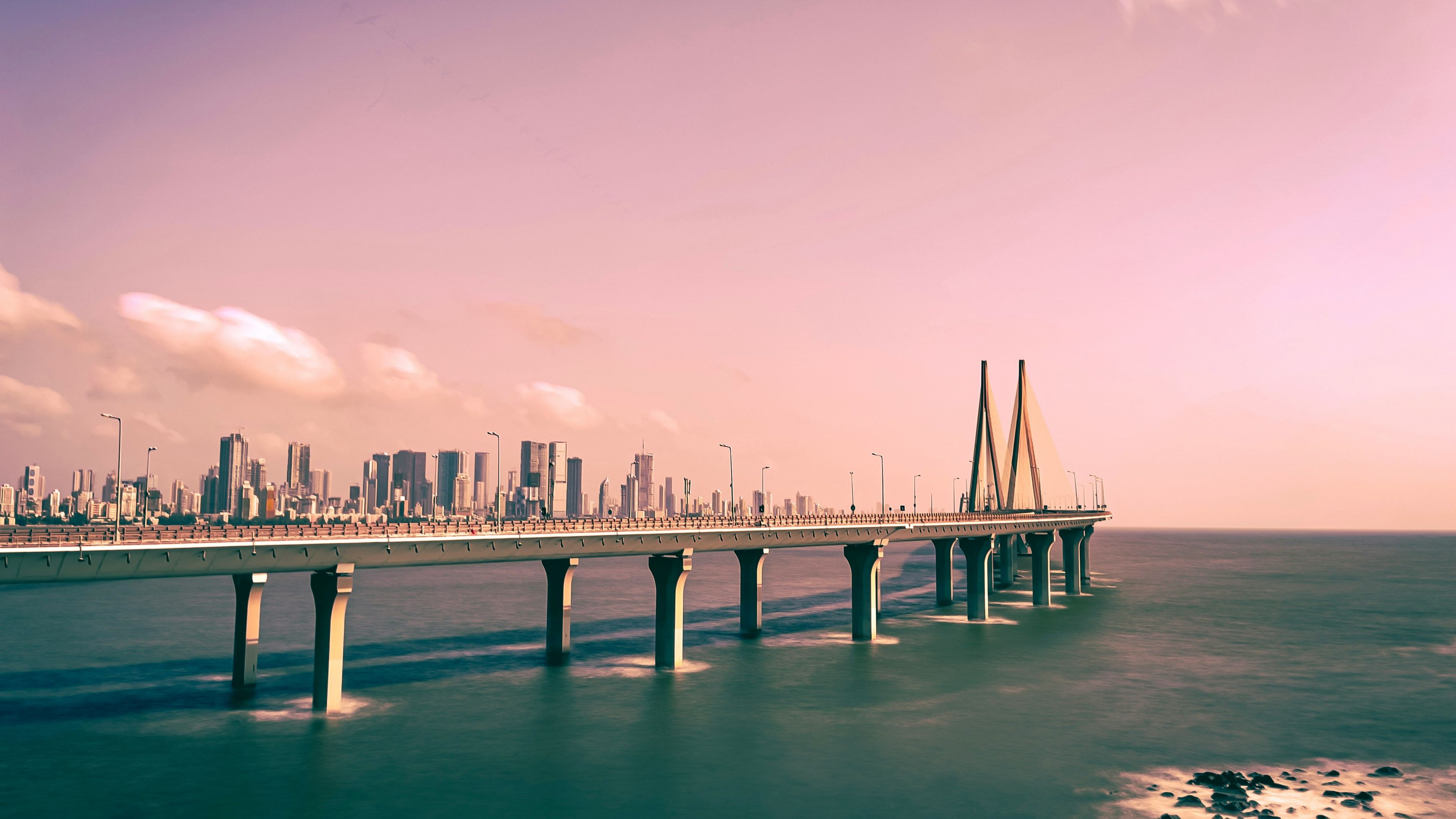 A bridge extends over the ocean towards a city skyline under a pink-tinted sky.