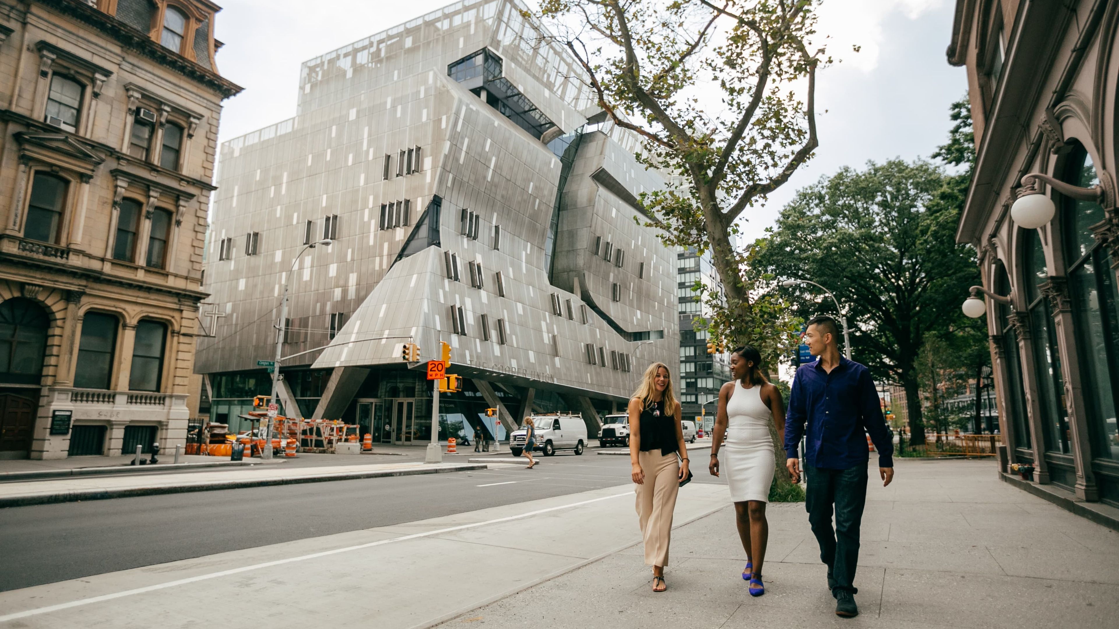 Three people walk on a city sidewalk near a modern, angular building.