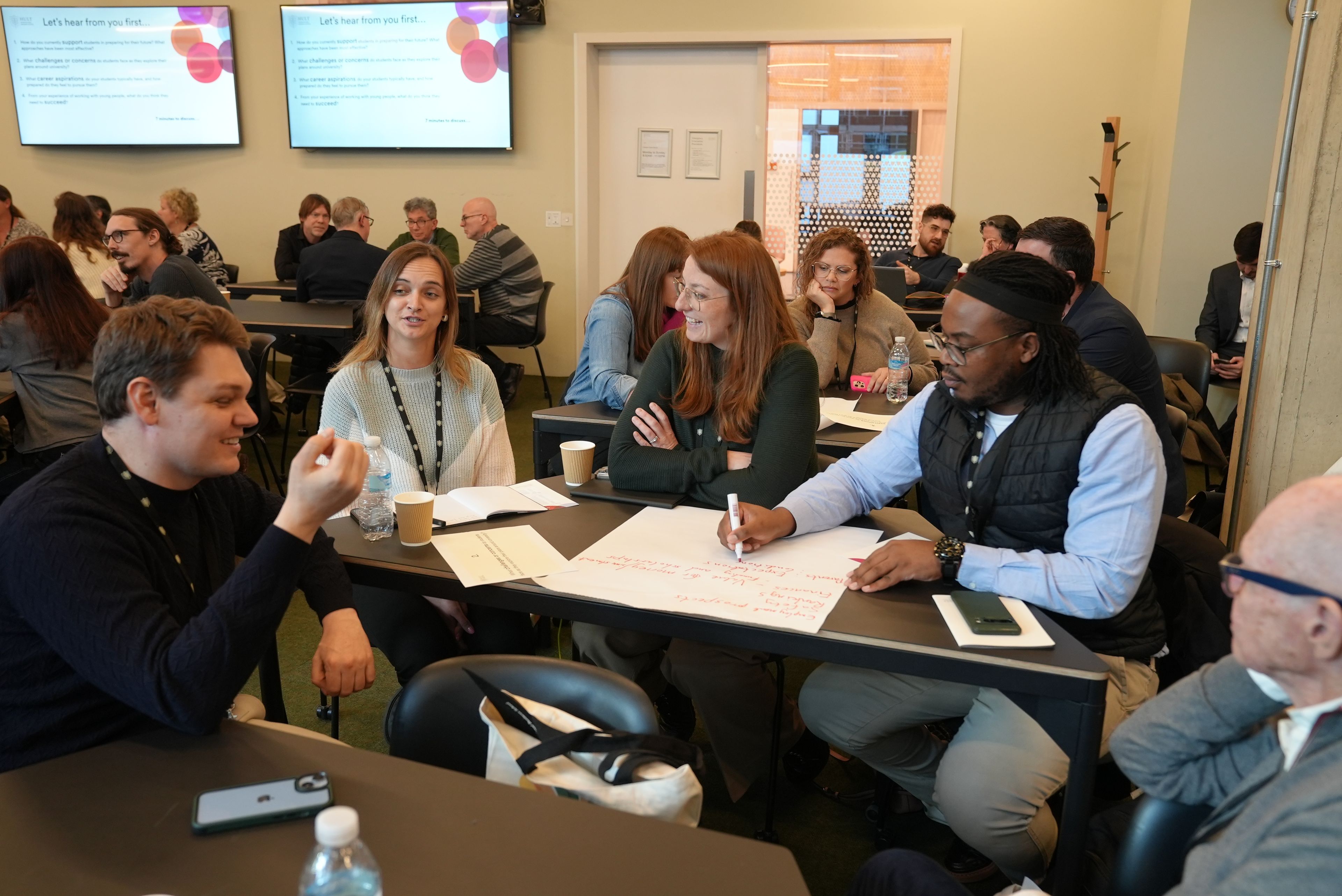 A group of people engaged in a discussion around a table, taking notes and sharing ideas in a conference room setting.