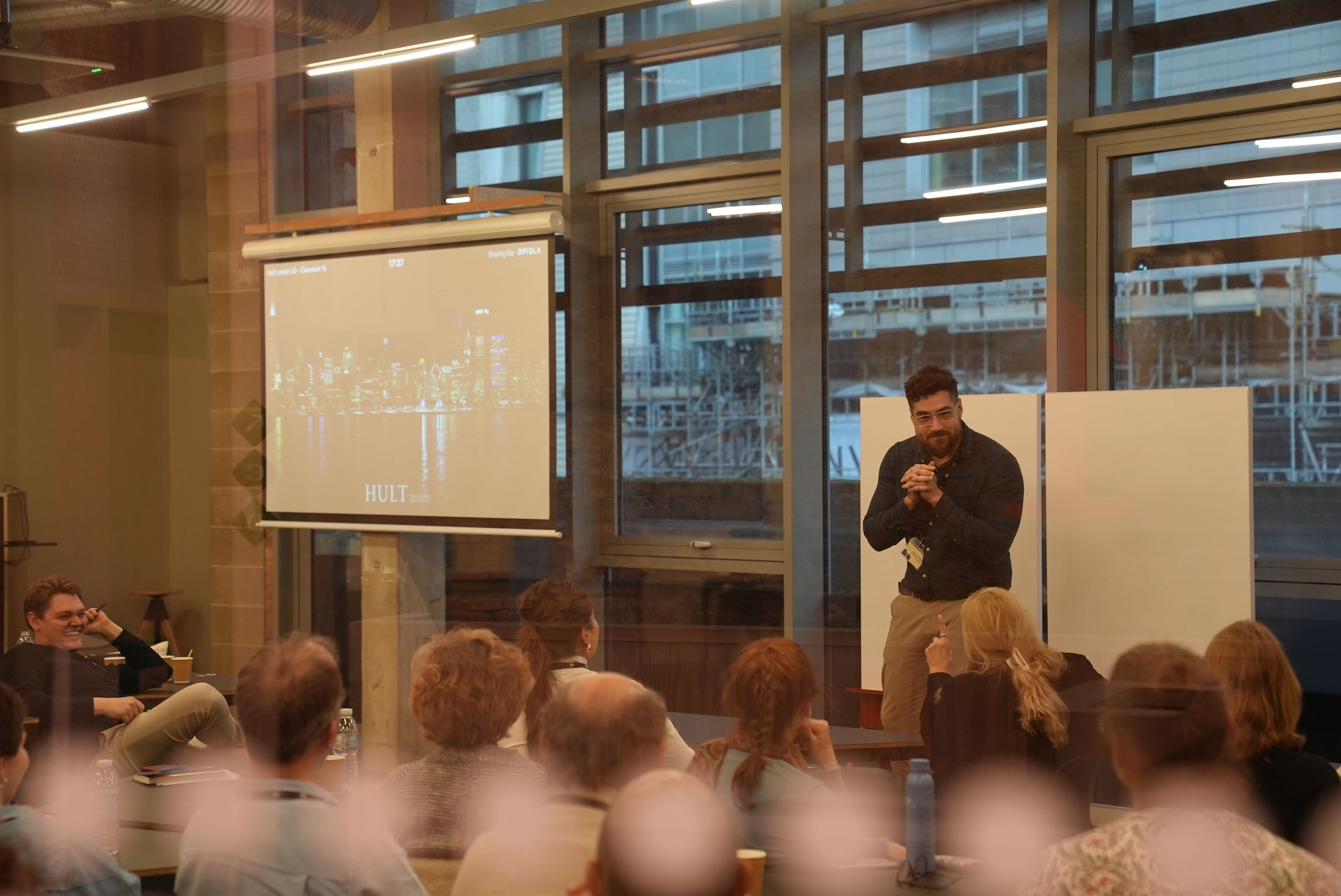 A man presents in a glass-walled room with a projected slide of a cityscape. Several people seated watch attentively.