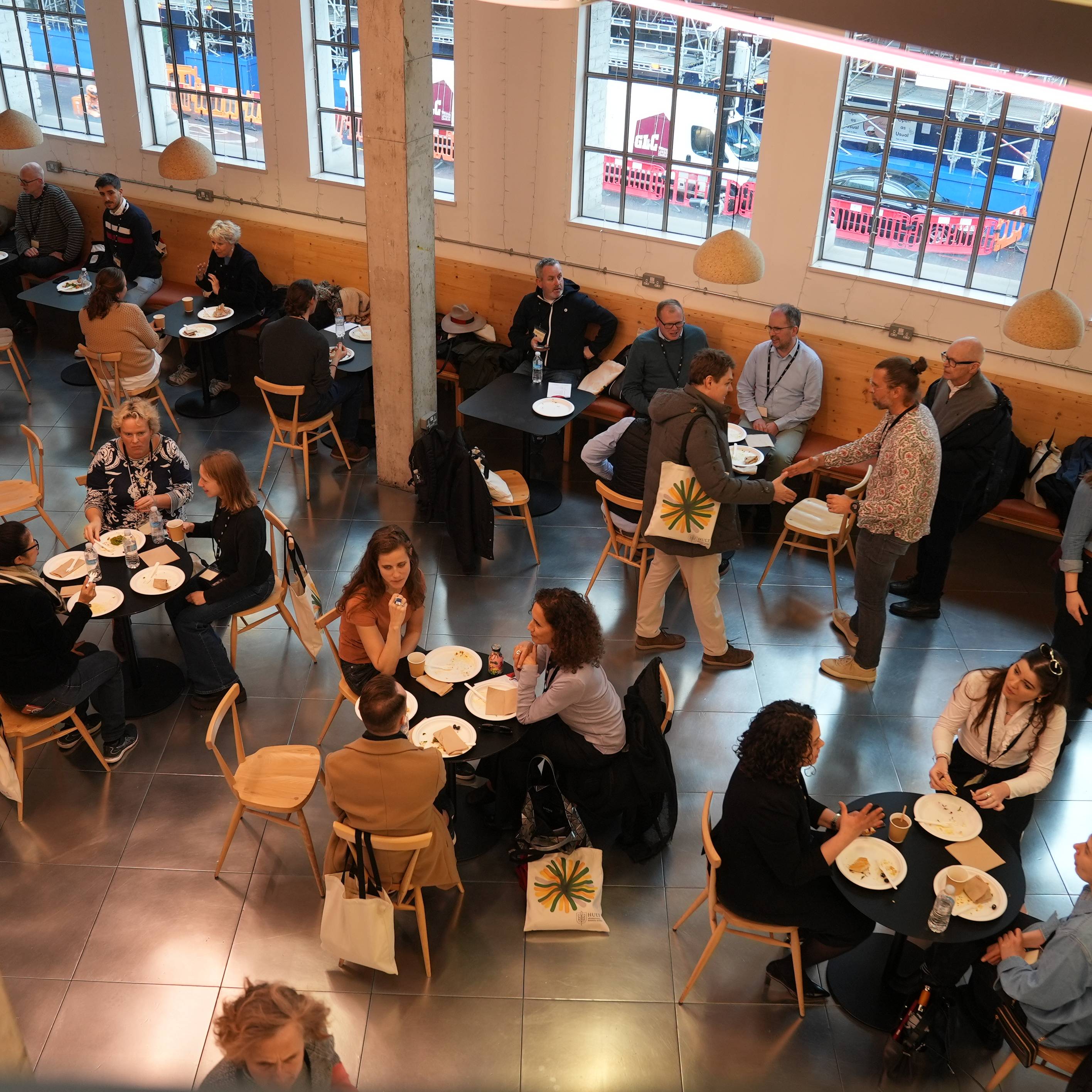 People seated at small tables in a bright, modern cafe, engaging in conversations and dining, with large windows in the background.