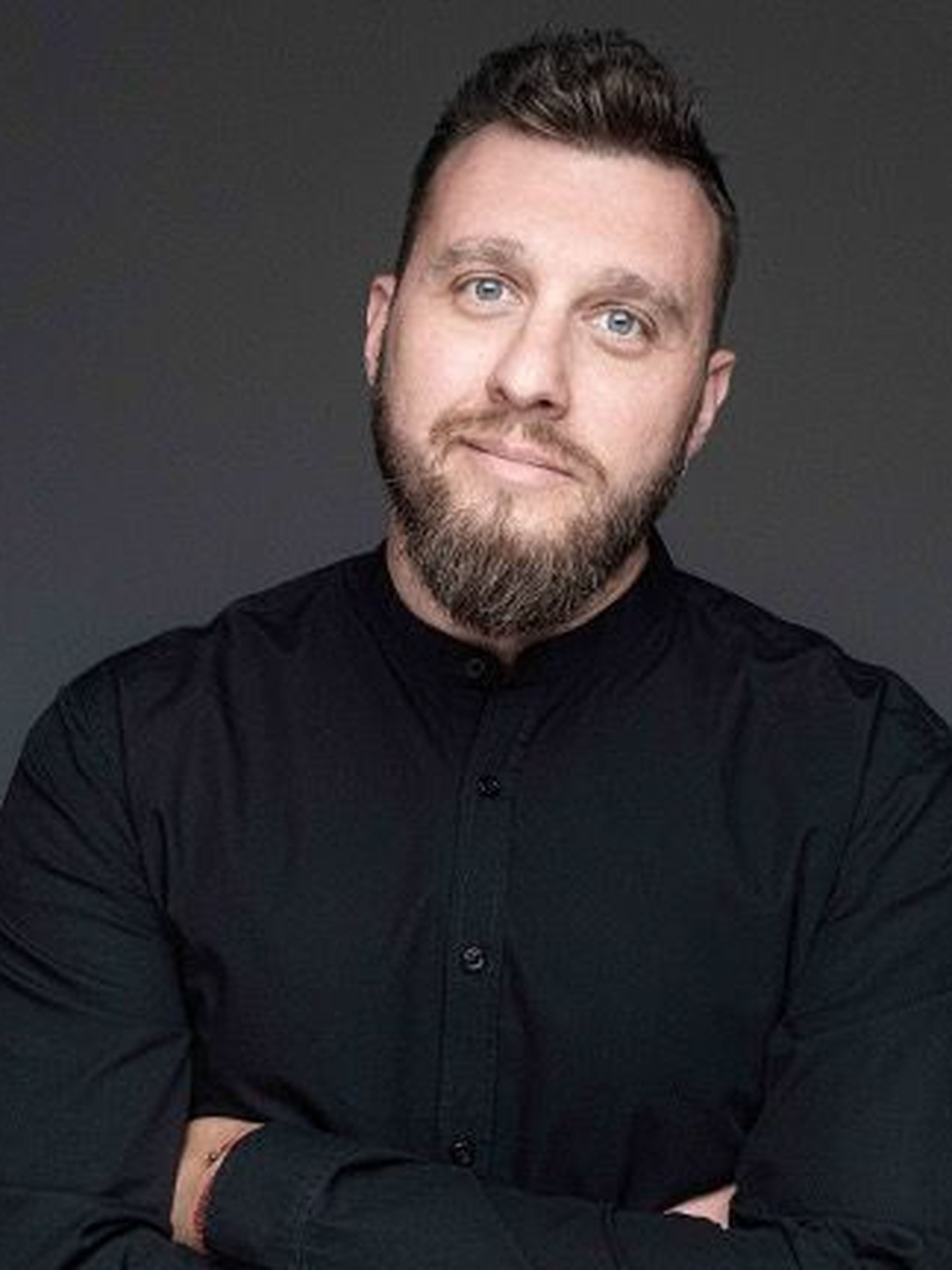 A man with a beard and a black shirt stands against a gray background.