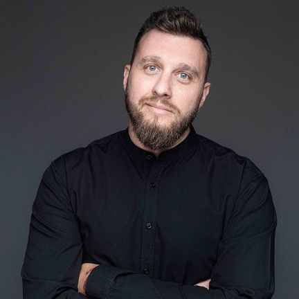 A man with a beard and a black shirt stands against a gray background.