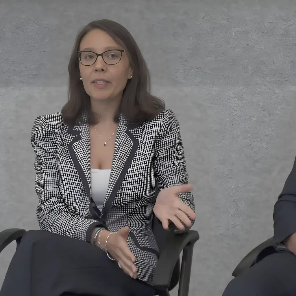 A woman in glasses sits and gestures while speaking, with a man seated beside her.
