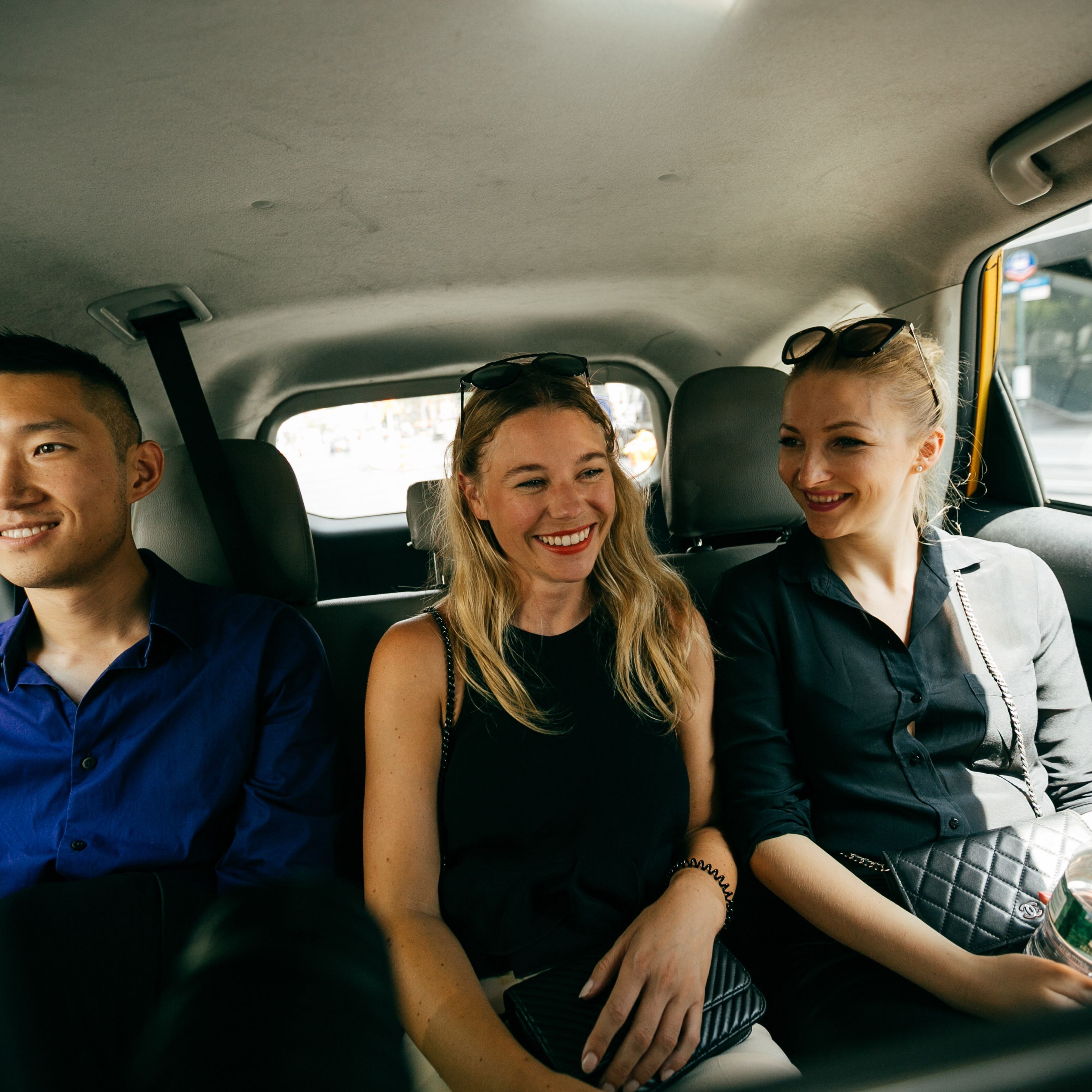 Three people smiling and chatting in the backseat of a car, with city buildings visible through the windows.