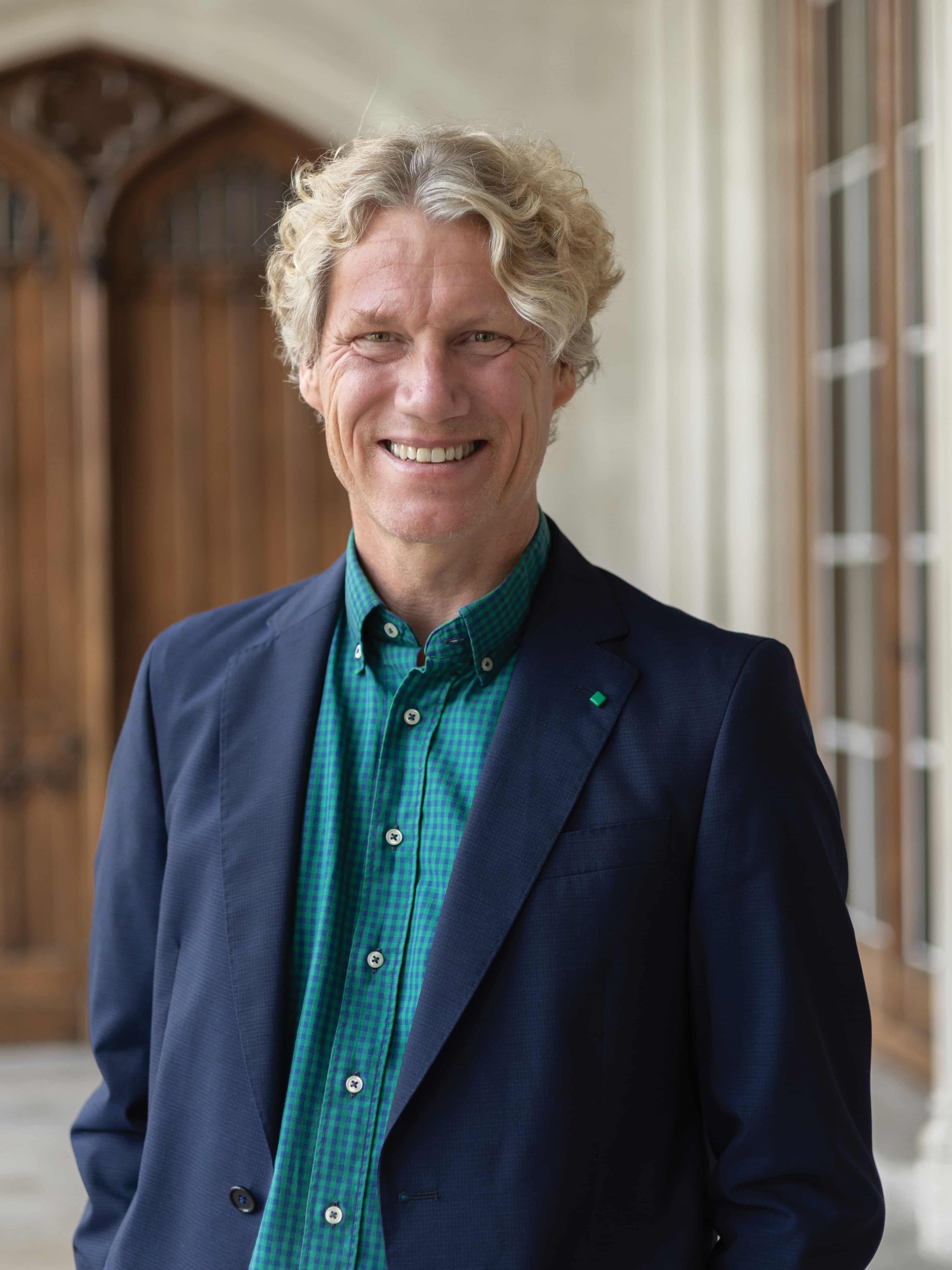 Smiling man with curly blonde hair wearing a blue jacket and green checkered shirt, standing in front of a wooden door.