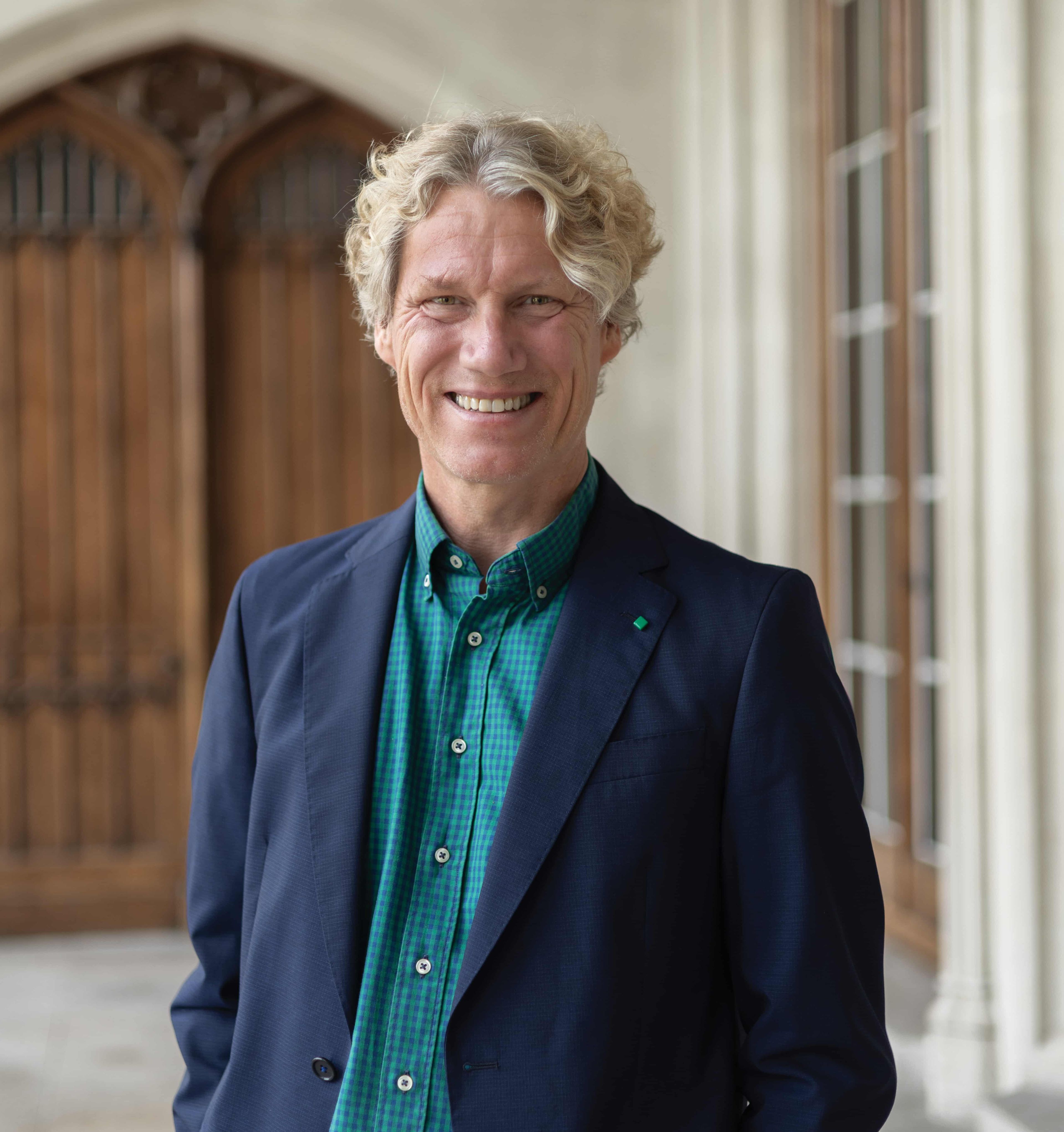 Smiling man with curly blonde hair wearing a blue jacket and green checkered shirt, standing in front of a wooden door.