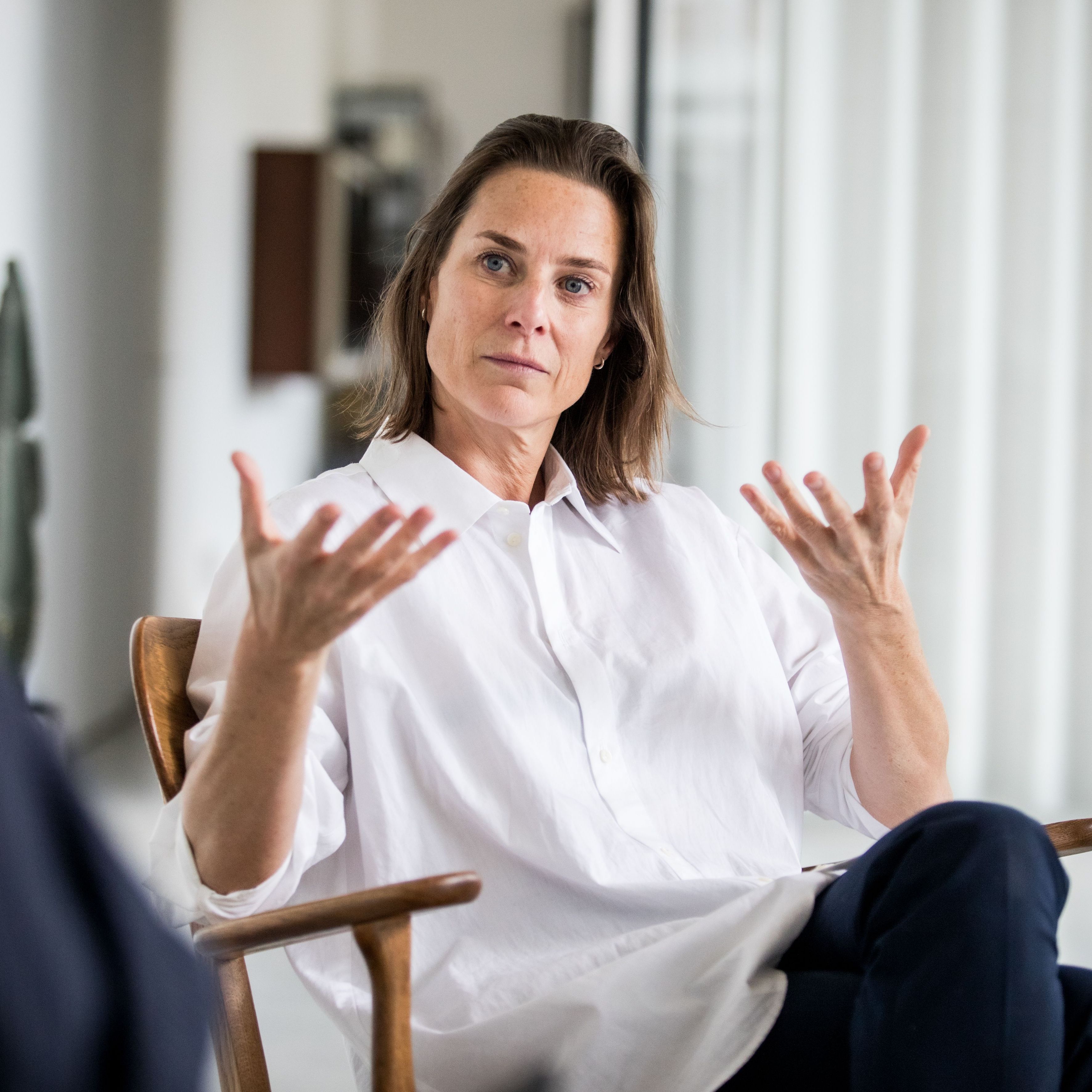Woman in a white shirt sits in a chair, gesturing with both hands while engaged in conversation in a bright room.