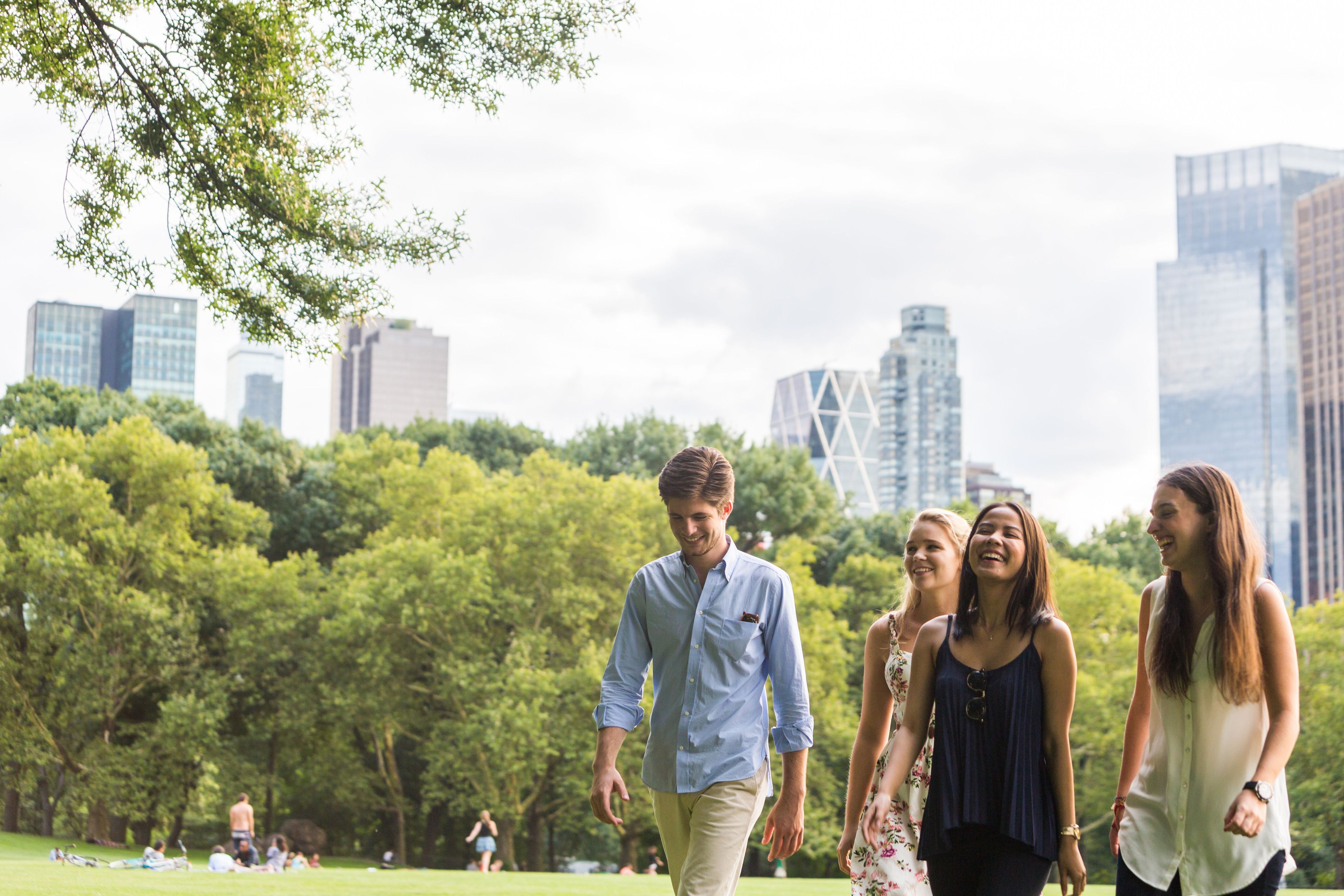 Four friends walking and laughing in a park with city skyline in the background.