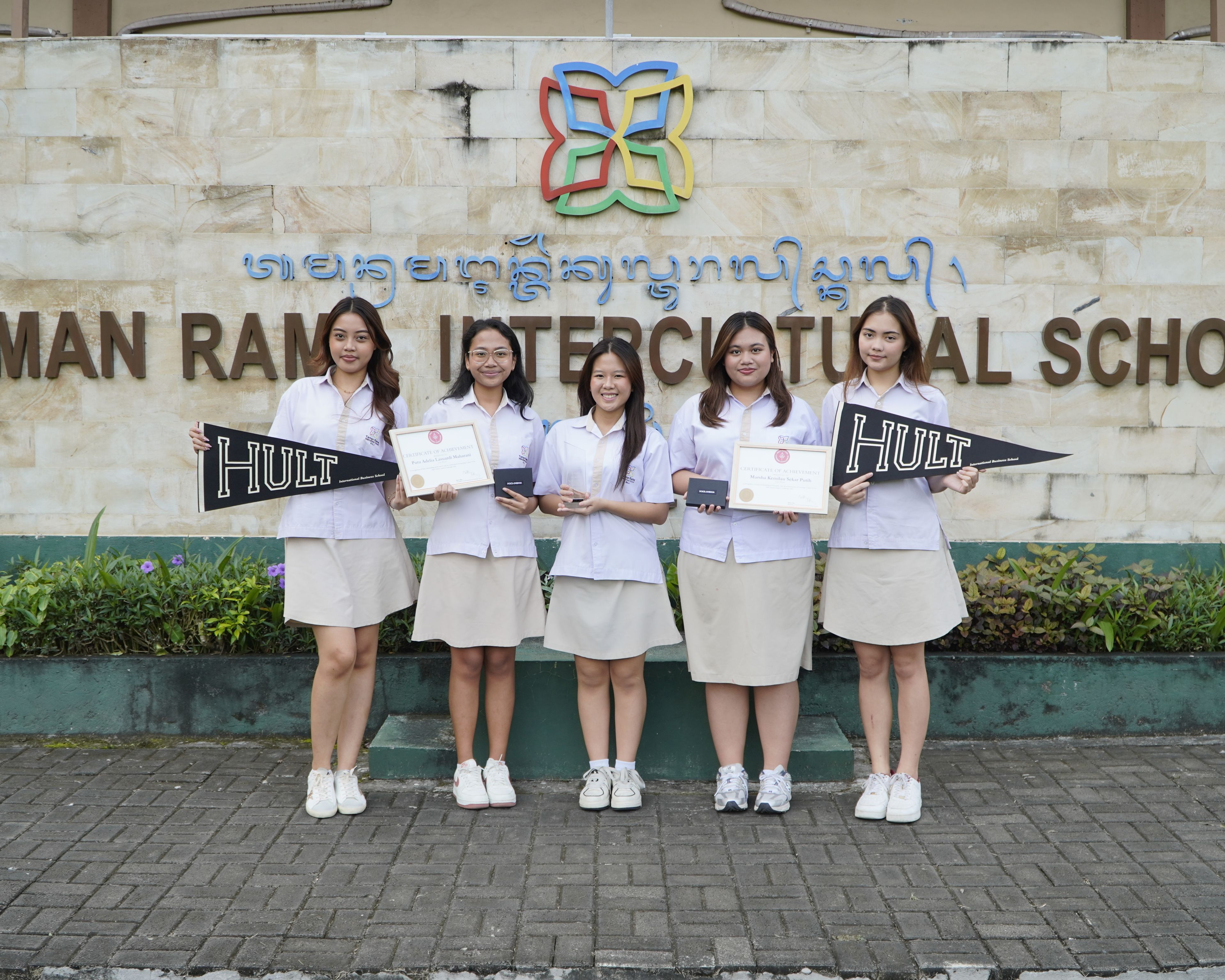 Five students in uniforms hold certificates and Hult flags in front of Taman Rama Intercultural School's sign.