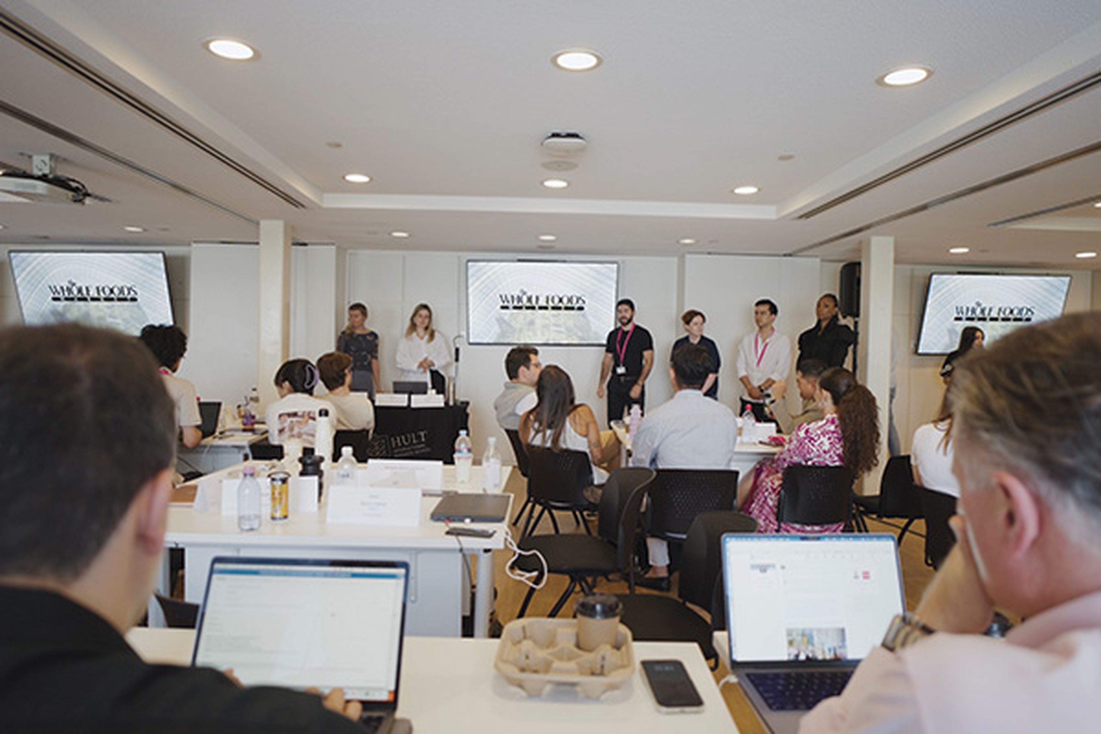 Students presenting in the classroom and supervisors seated at the back.