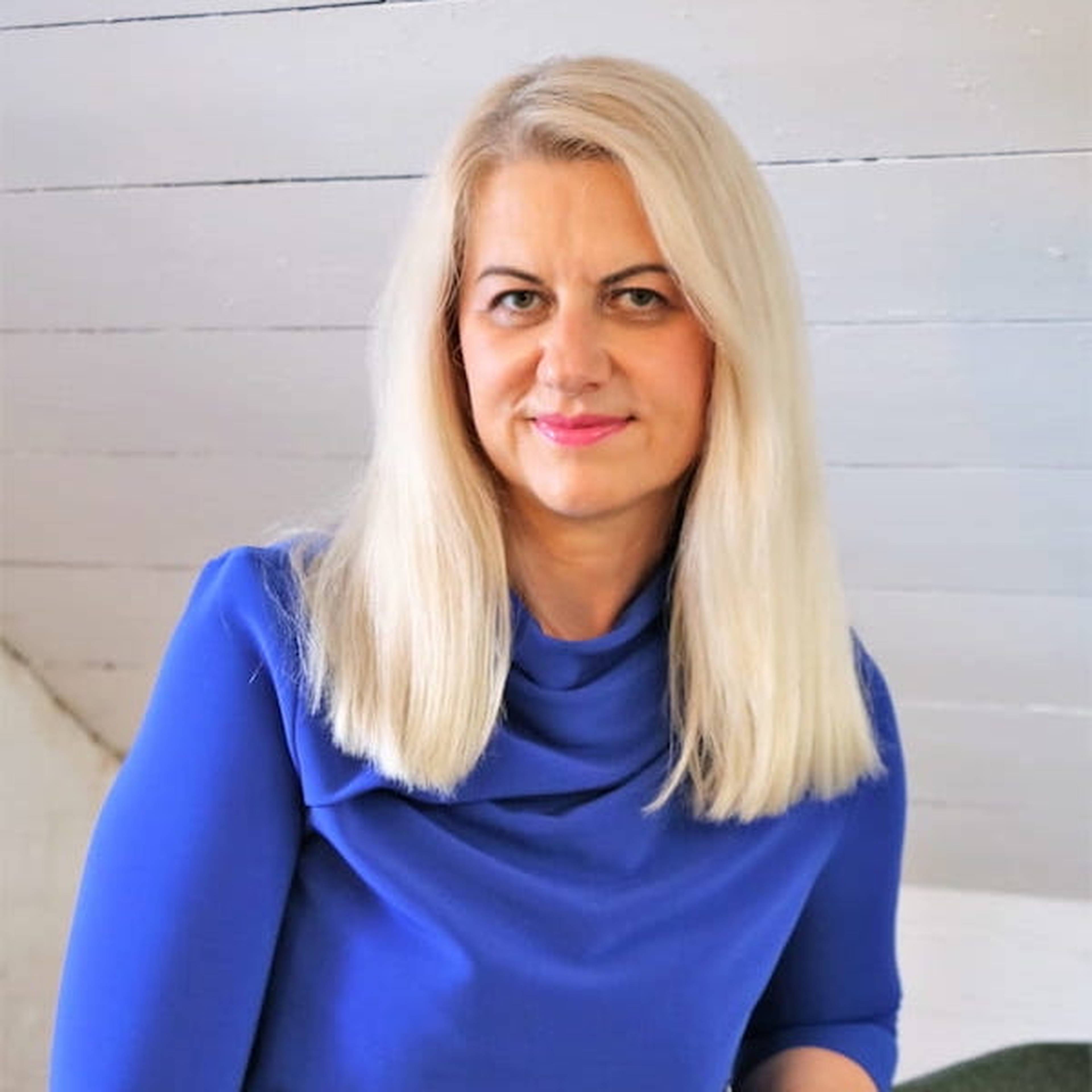 Woman with long blonde hair wearing a blue dress, sitting indoors against a white wooden wall.