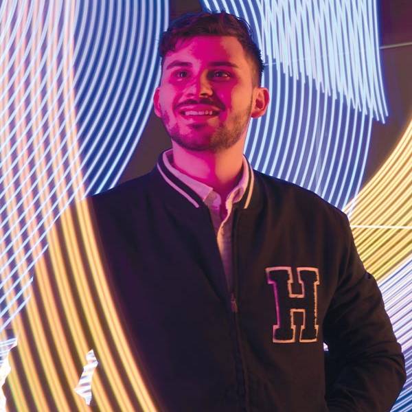 Smiling man in a jacket with stands in front of vibrant, swirling neon light patterns in blue and yellow.