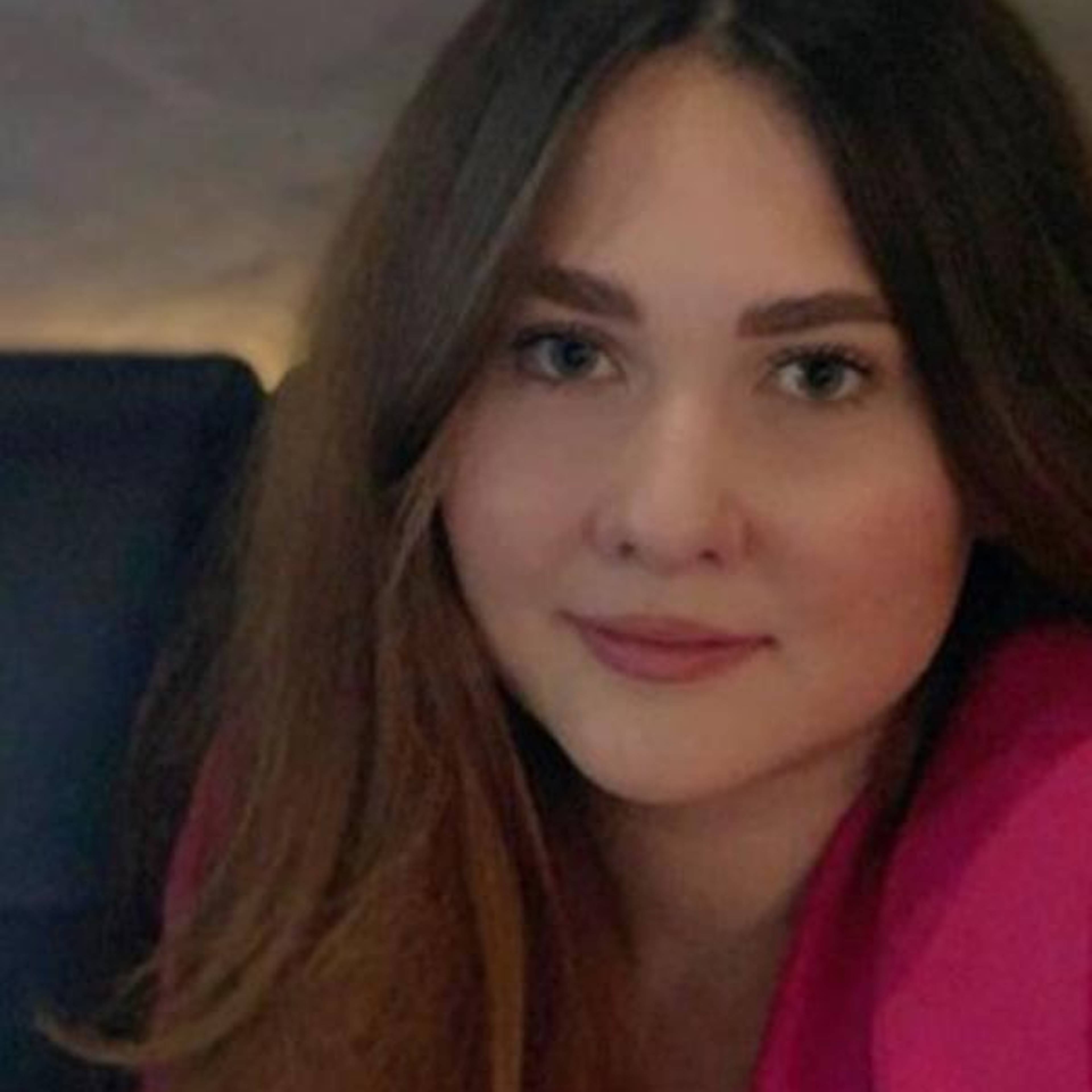 A woman with long hair and a pink top is smiling softly at the camera.