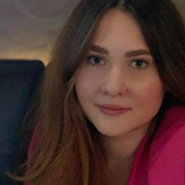 A woman with long hair and a pink top is smiling softly at the camera.