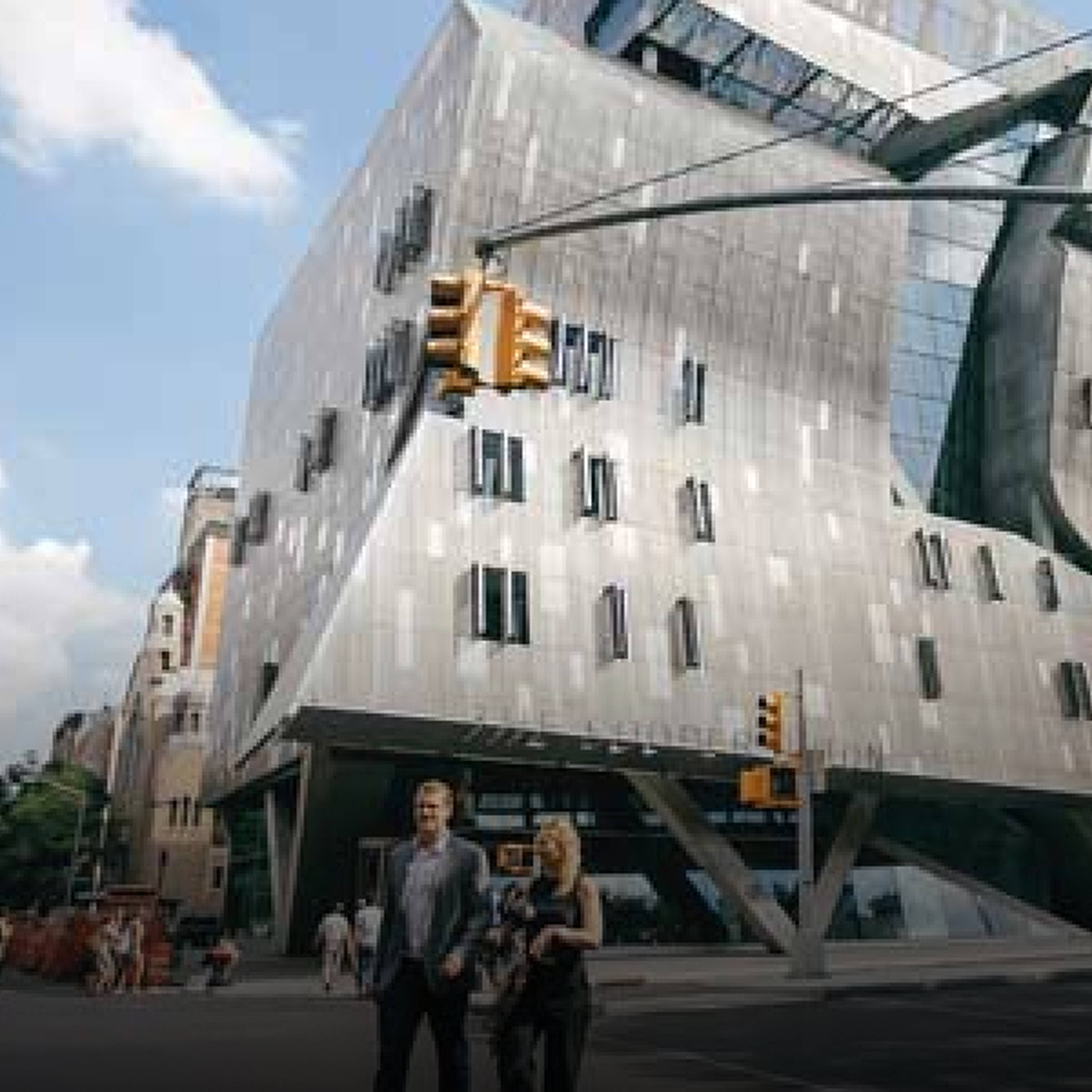 People walk near a modern, angular building under a bright blue sky with clouds.