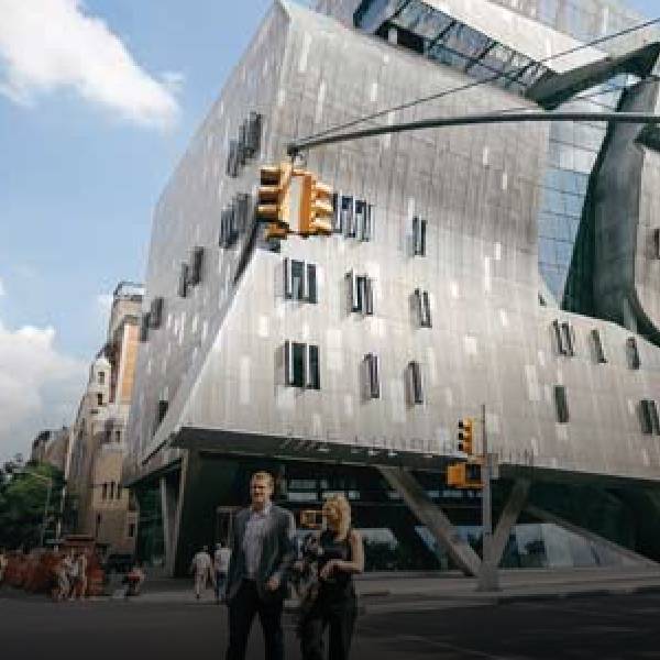 People walk near a modern, angular building under a bright blue sky with clouds.